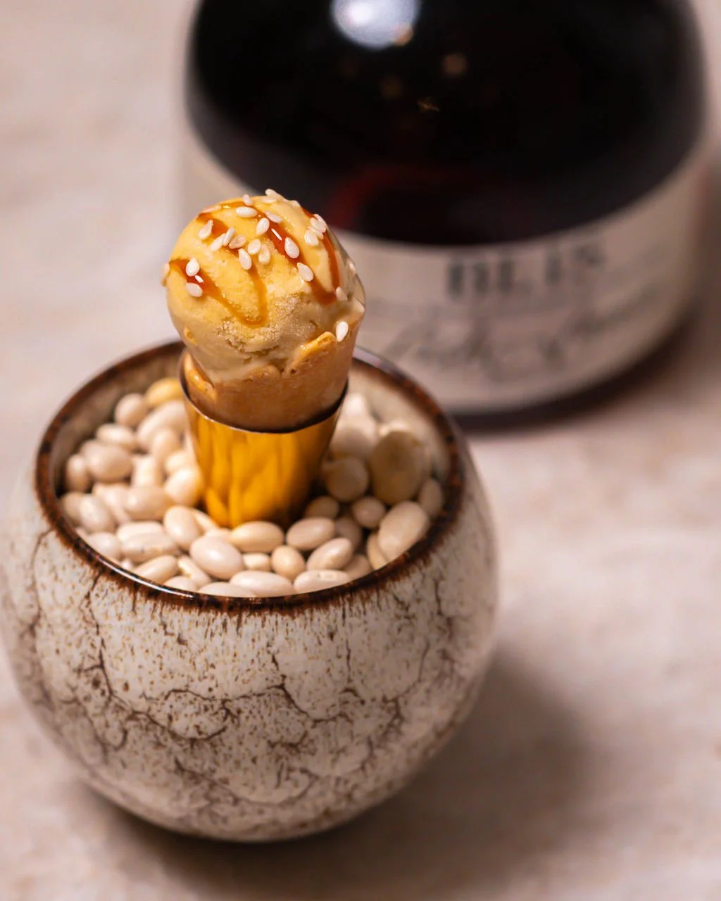 Ice cream cone with caramel drizzle and white sprinkles in a decorative ceramic holder filled with white beans and a black bottle in the background.