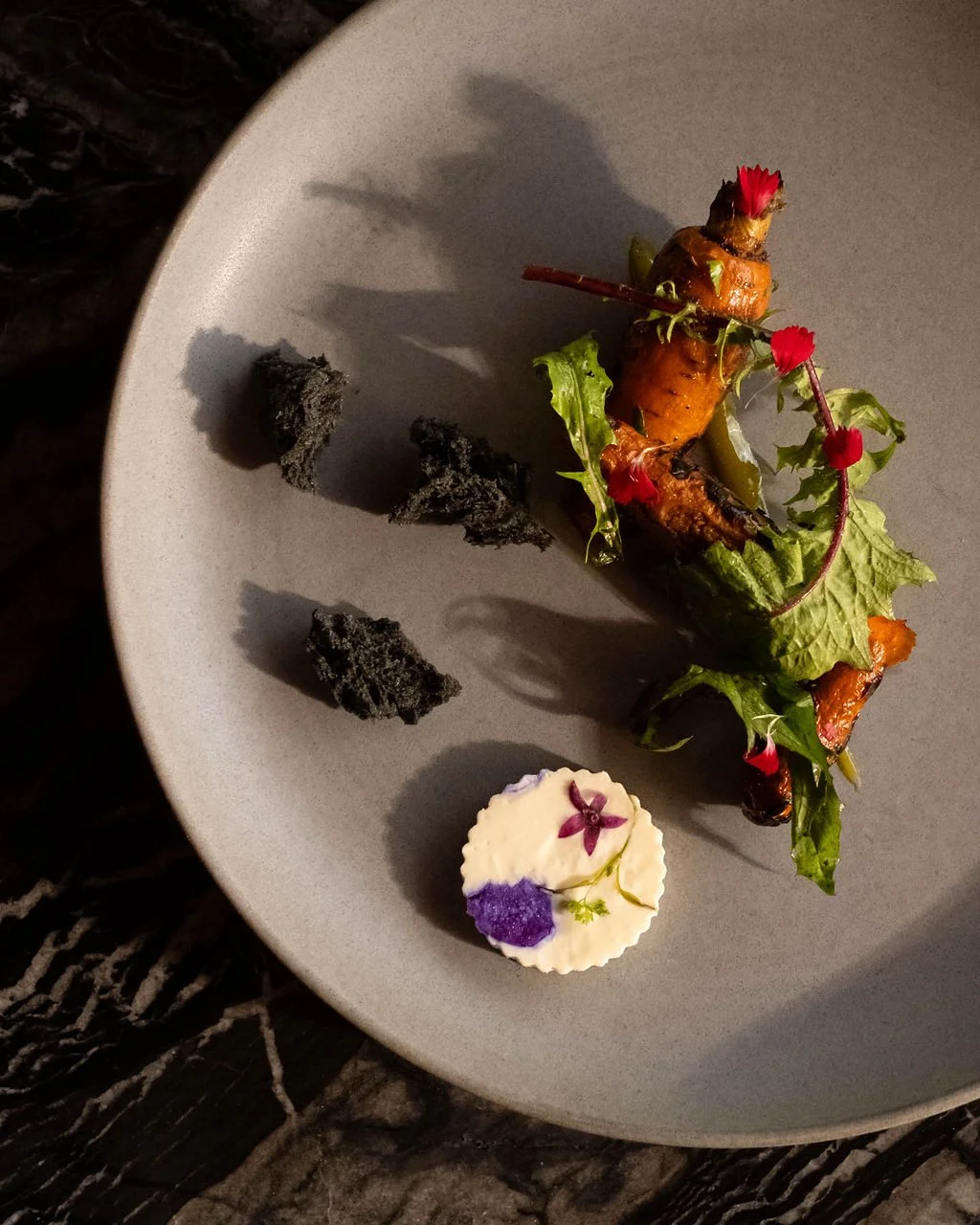 Elegant plated dish featuring roasted root vegetables with greens and edible flowers, accompanied by black garnishes and a dollop of creamy sauce with purple and green accents.