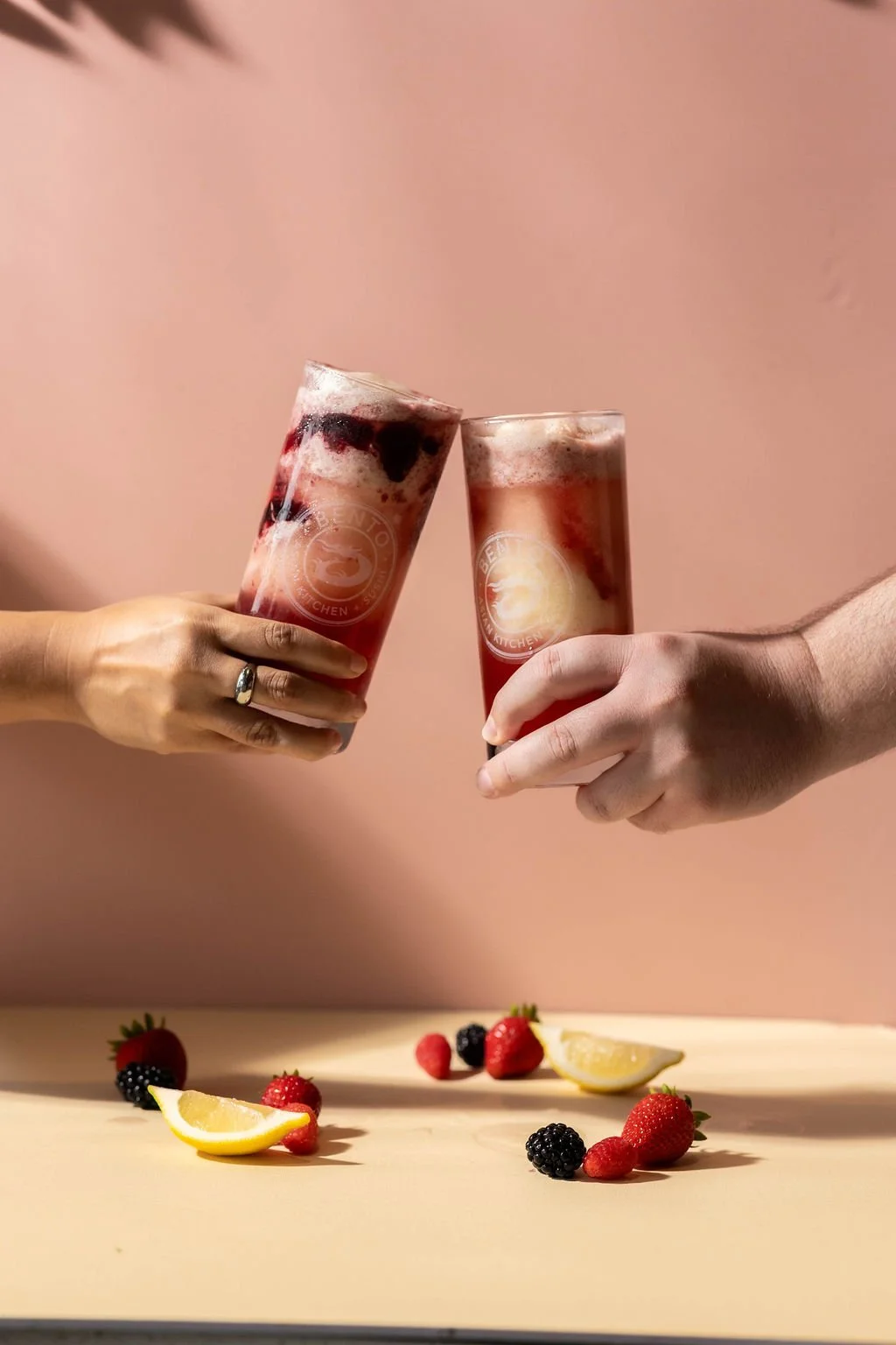 Two people clinking glasses of berry beverages with ice, on a light-colored surface with strawberries, blackberries, and lemon wedges scattered below. The background is pink.