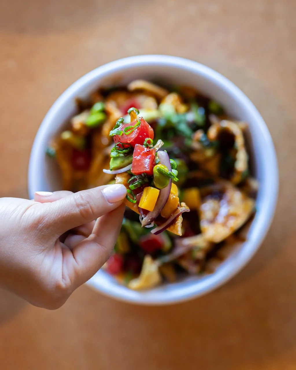 Close-up of a hand holding a spoonful of vegetable stir-fry above a bowl filled with similar stir-fried vegetables and chicken, with a wooden surface background.