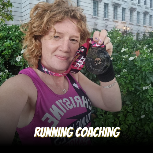 A woman with curly red hair in a purple running tank top holding a medal with a pink ribbon, standing outdoors with green bushes and a building in the background.