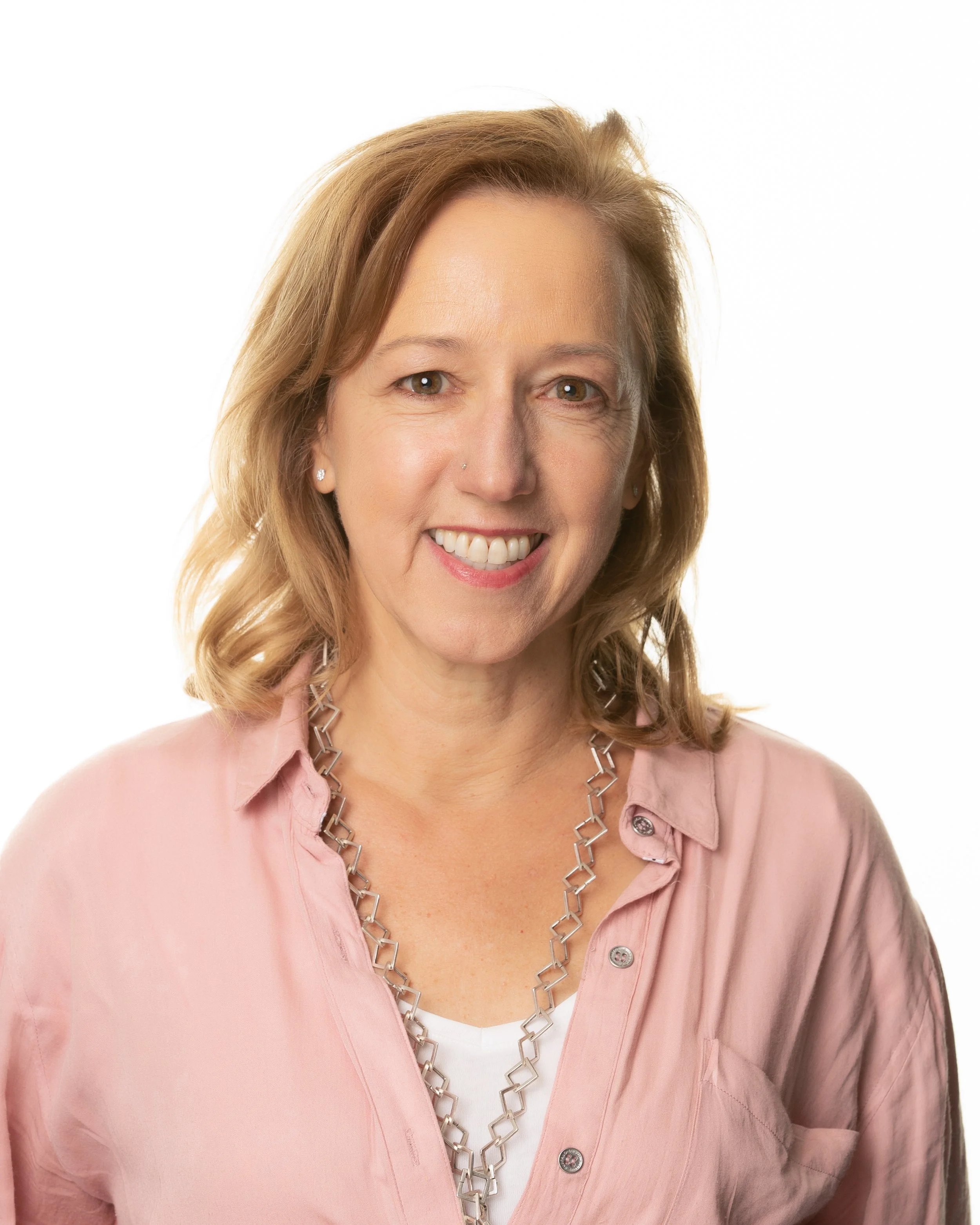 A woman with shoulder-length blonde hair smiling, wearing a pink shirt and a silver necklace, against a white background.