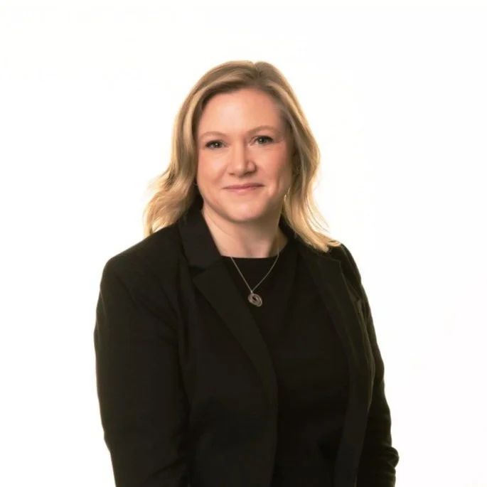 Portrait of a blonde woman wearing a black blazer and a necklace, smiling at the camera against a white background.