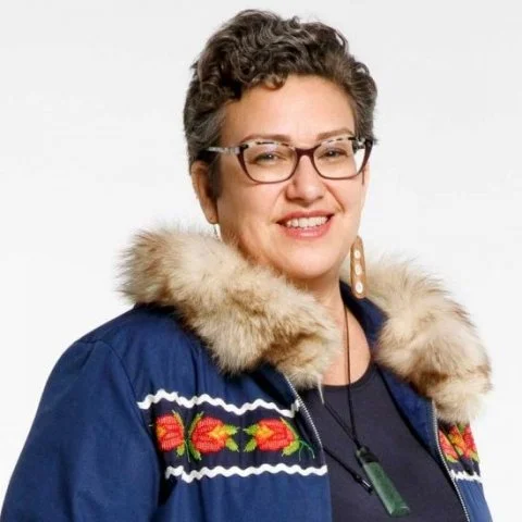Portrait of a smiling woman with short curly hair and glasses, wearing a blue jacket with fur trim and colorful embroidery.