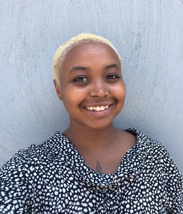 Close-up of a smiling woman with short blonde hair, wearing a black and white patterned top, standing against a plain gray wall.