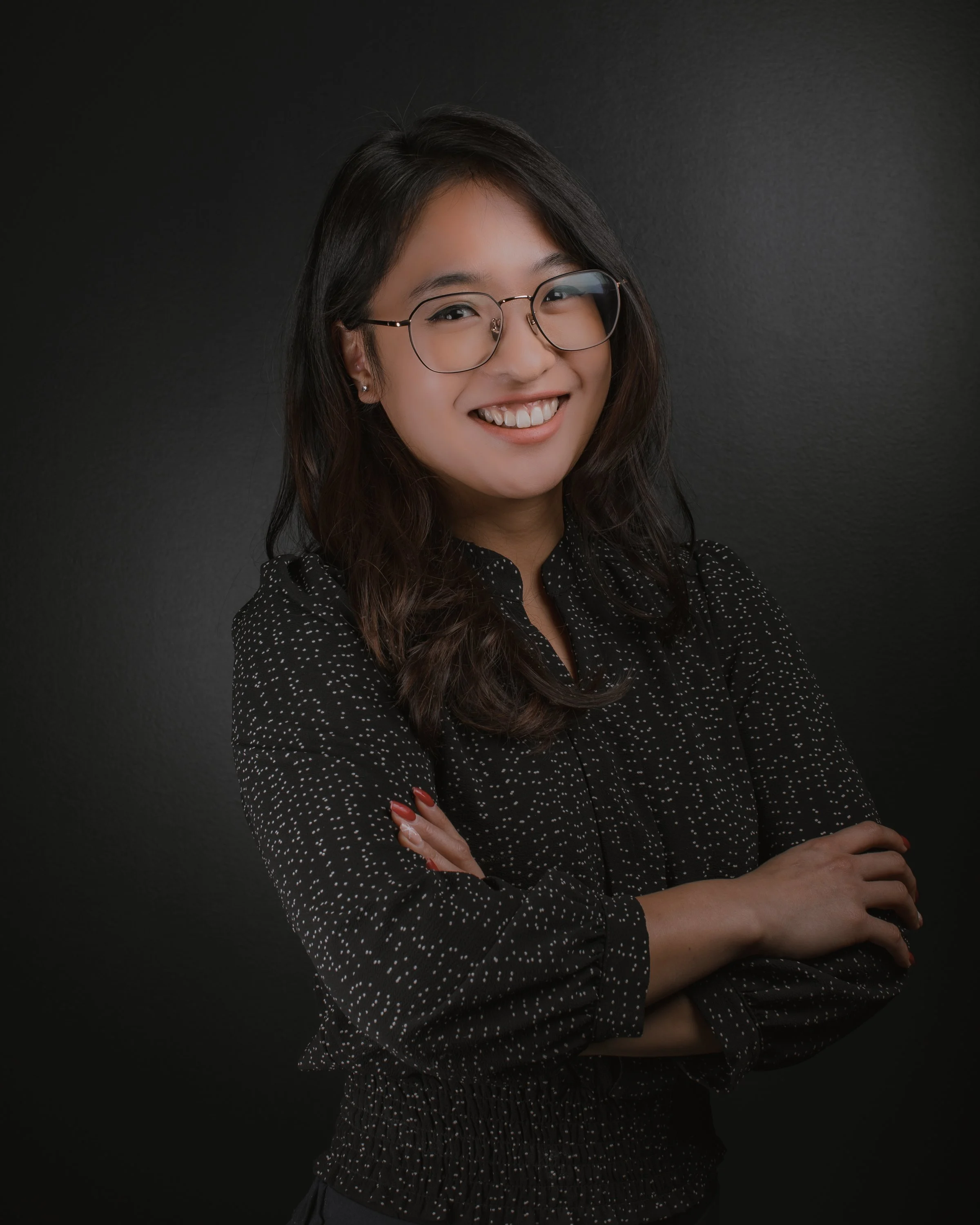 A young woman with glasses, smiling, wearing a black dress with white dots and crossing her arms.