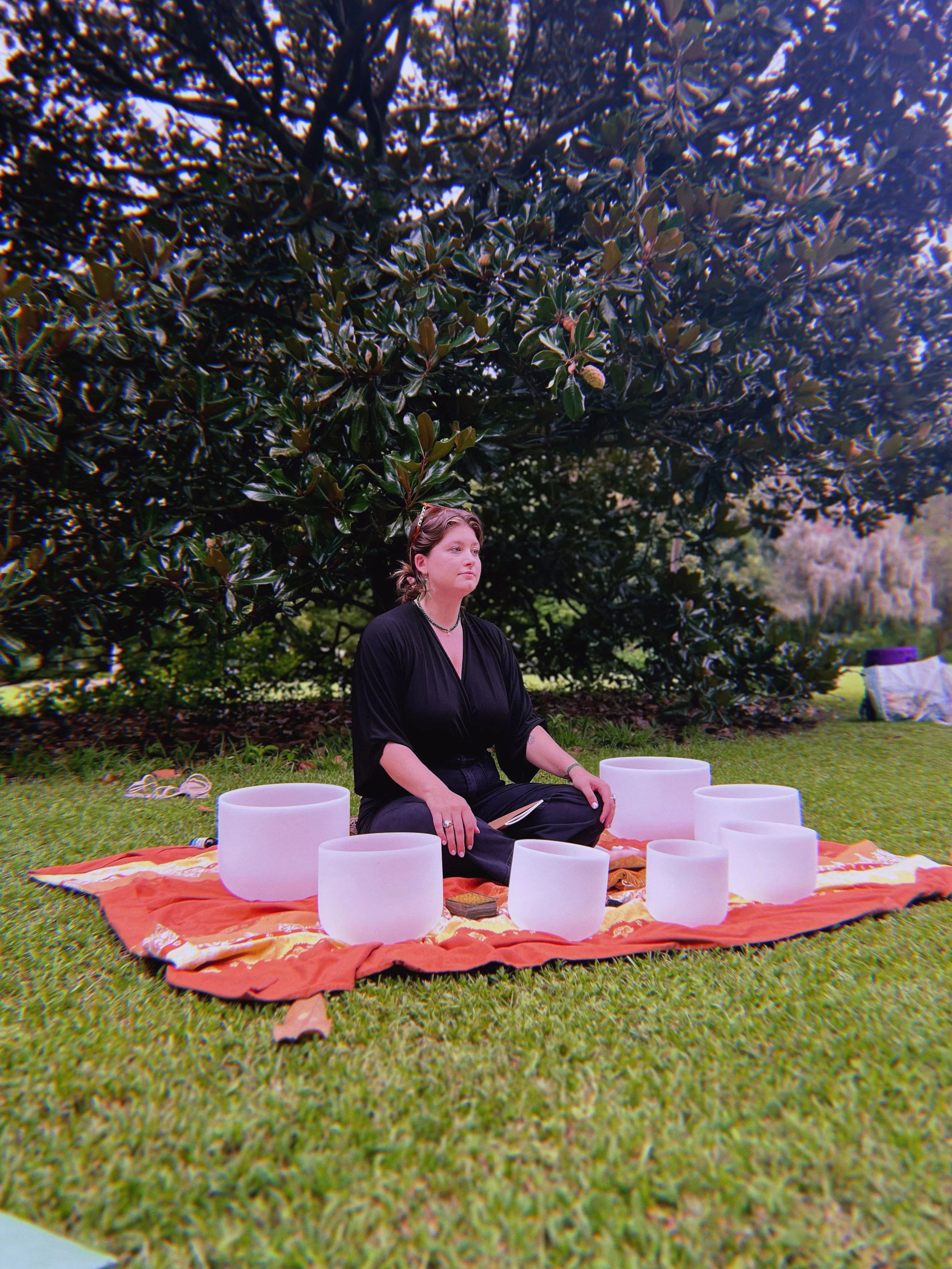 Person sitting cross-legged on an orange and gold blanket on grass, surrounded by white crystal singing bowls outdoors under a large leafy tree.