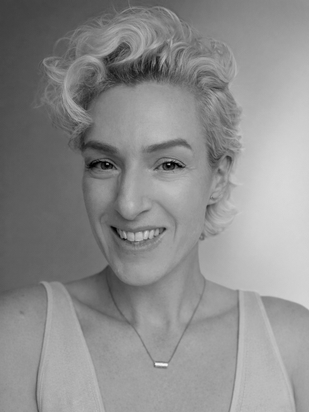 Black and white portrait of a smiling woman with short, curly hair wearing a sleeveless top and a necklace.