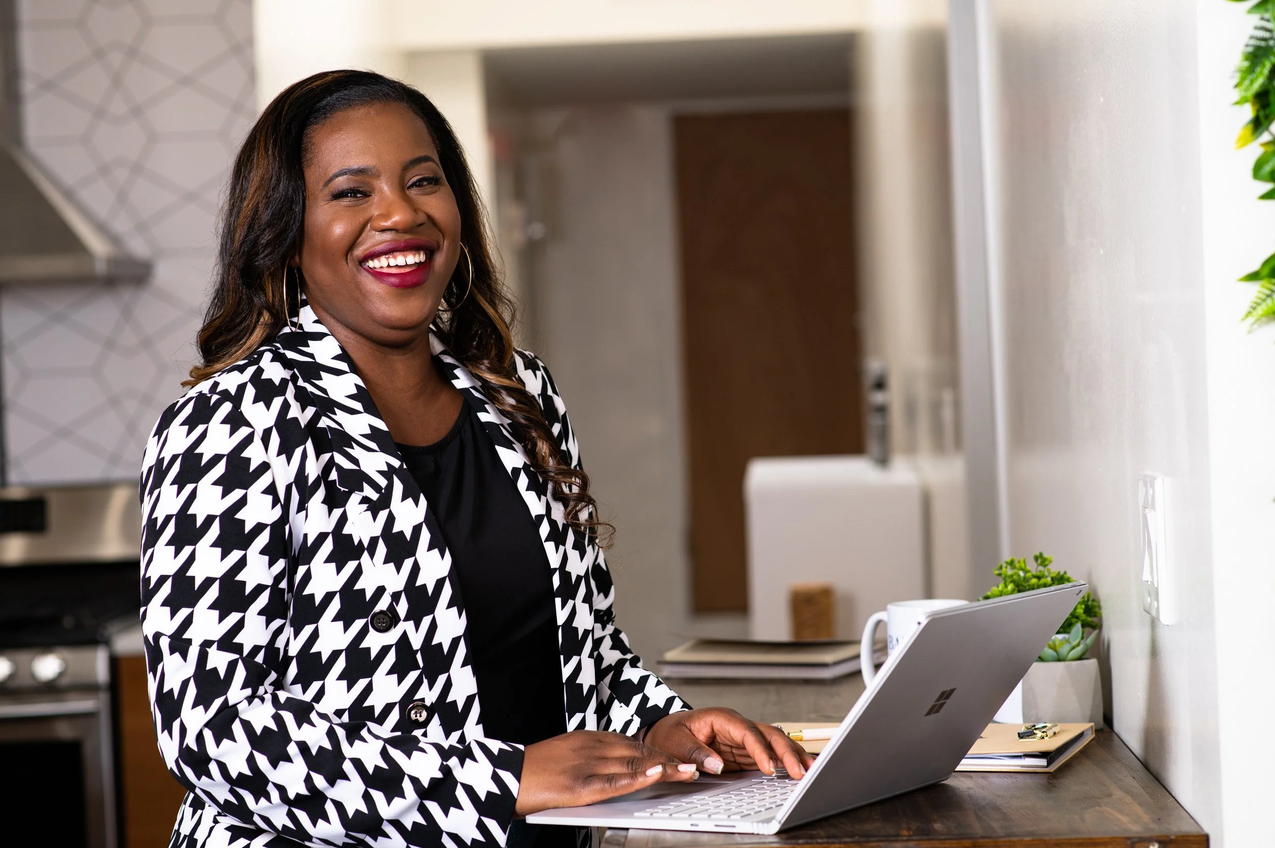 Corporate Event Planner Tahirah wears a black and white houndstooth blazer while smiling at the camera and typing at her laptop.