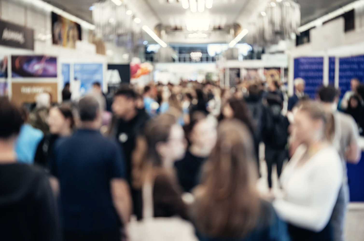 Blurred crowd of people walking inside a busy exhibition or conference hall with various booths and displays.