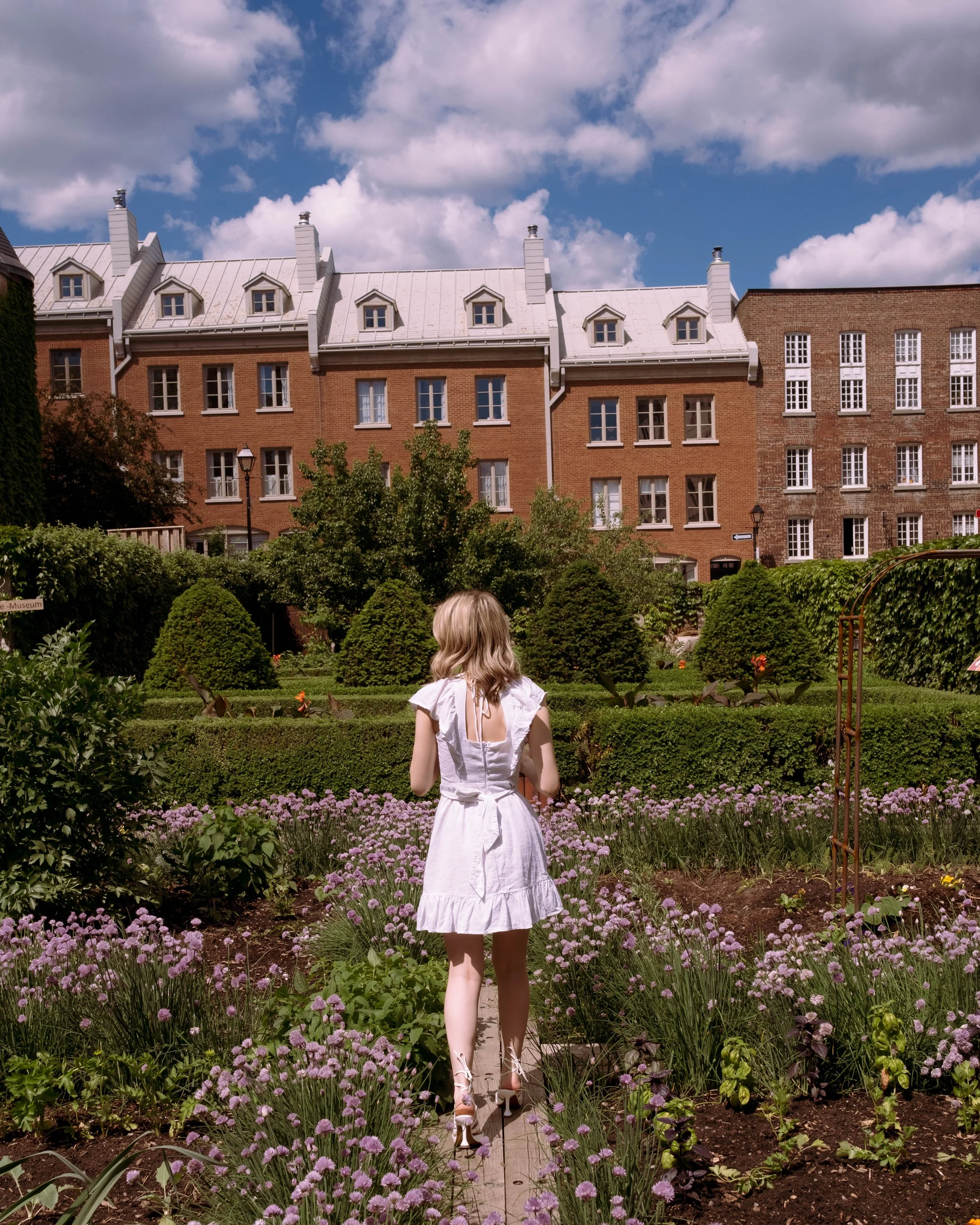 A woman in a white dress walking on a garden path surrounded by purple flowers, with brick buildings and a partly cloudy sky in the background.