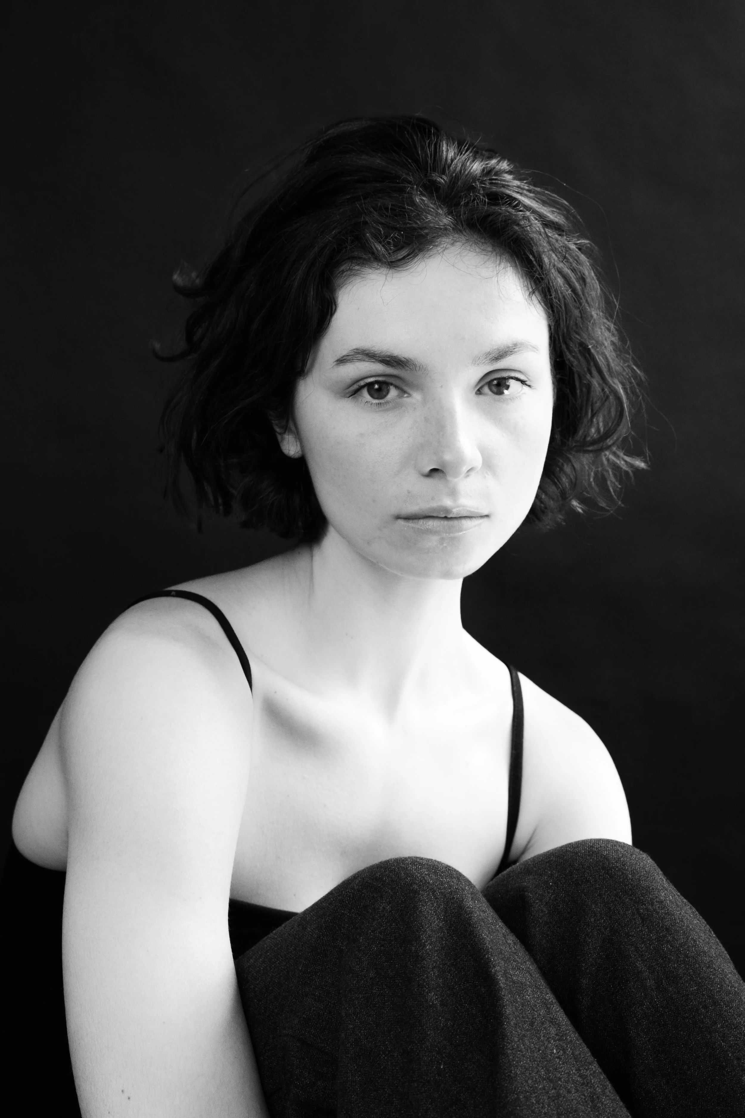 A black-and-white portrait of a woman with short wavy hair, wearing a spaghetti strap top, sitting against a dark background, looking directly at the camera.