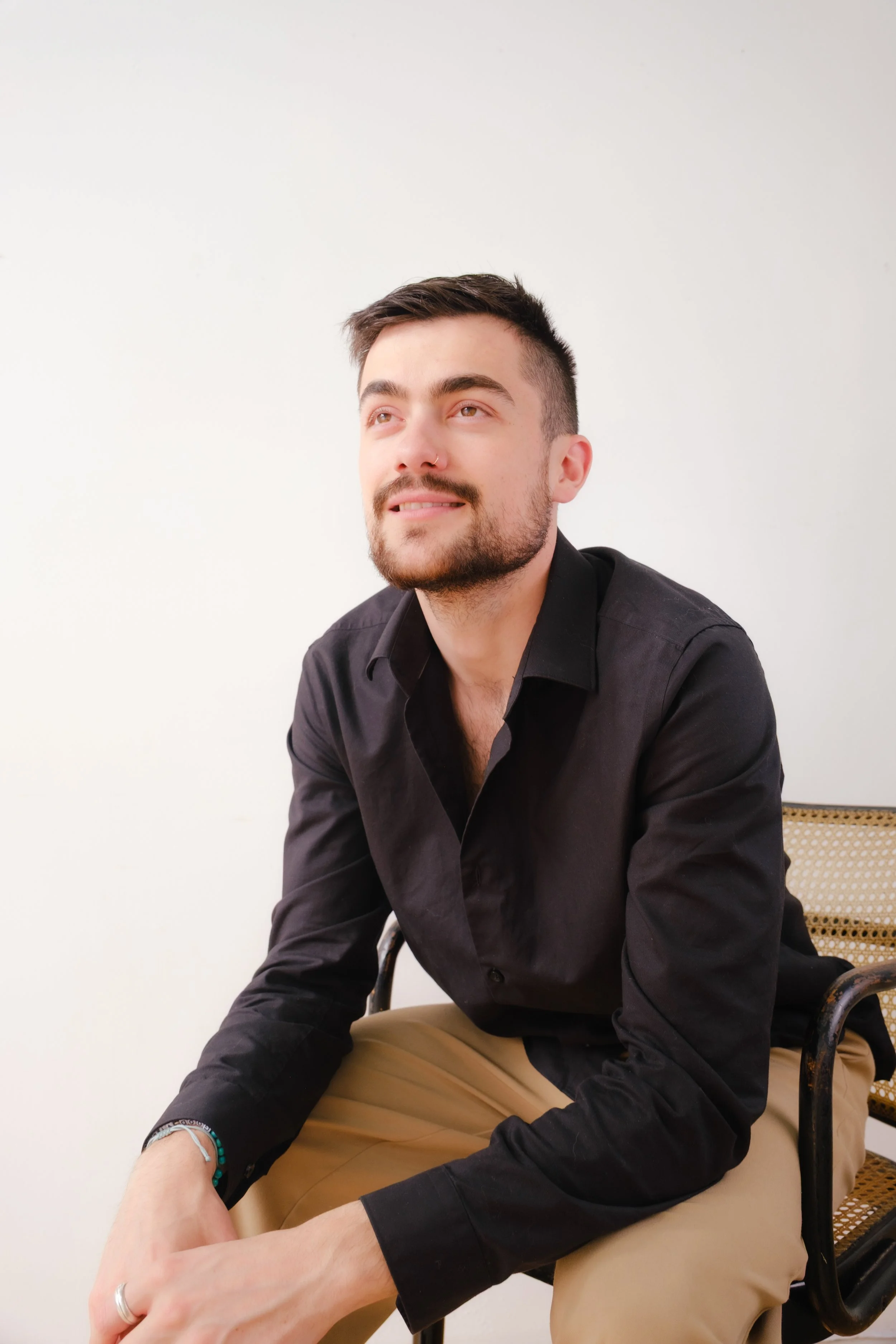 A young man with short dark hair, a beard, and a nose piercing sits on a chair against a plain white wall, wearing a black shirt and tan pants, looking slightly upward with a smile.