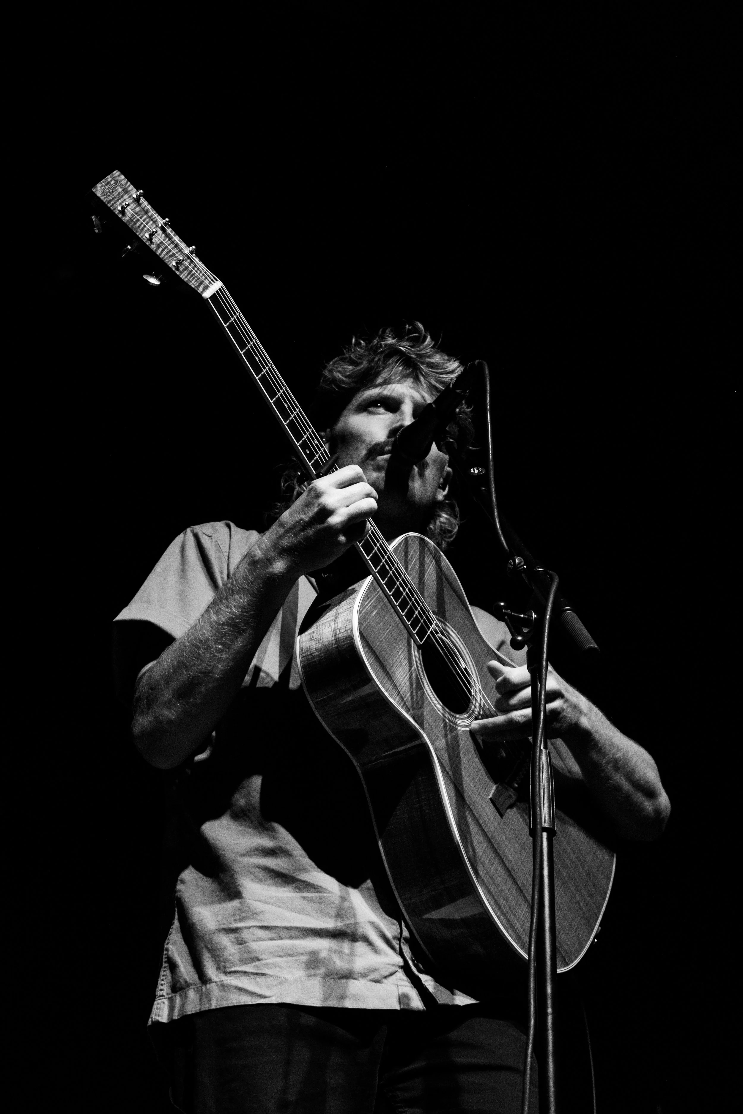 A man with long hair playing an acoustic guitar on stage, singing into a microphone. The image is in black and white.