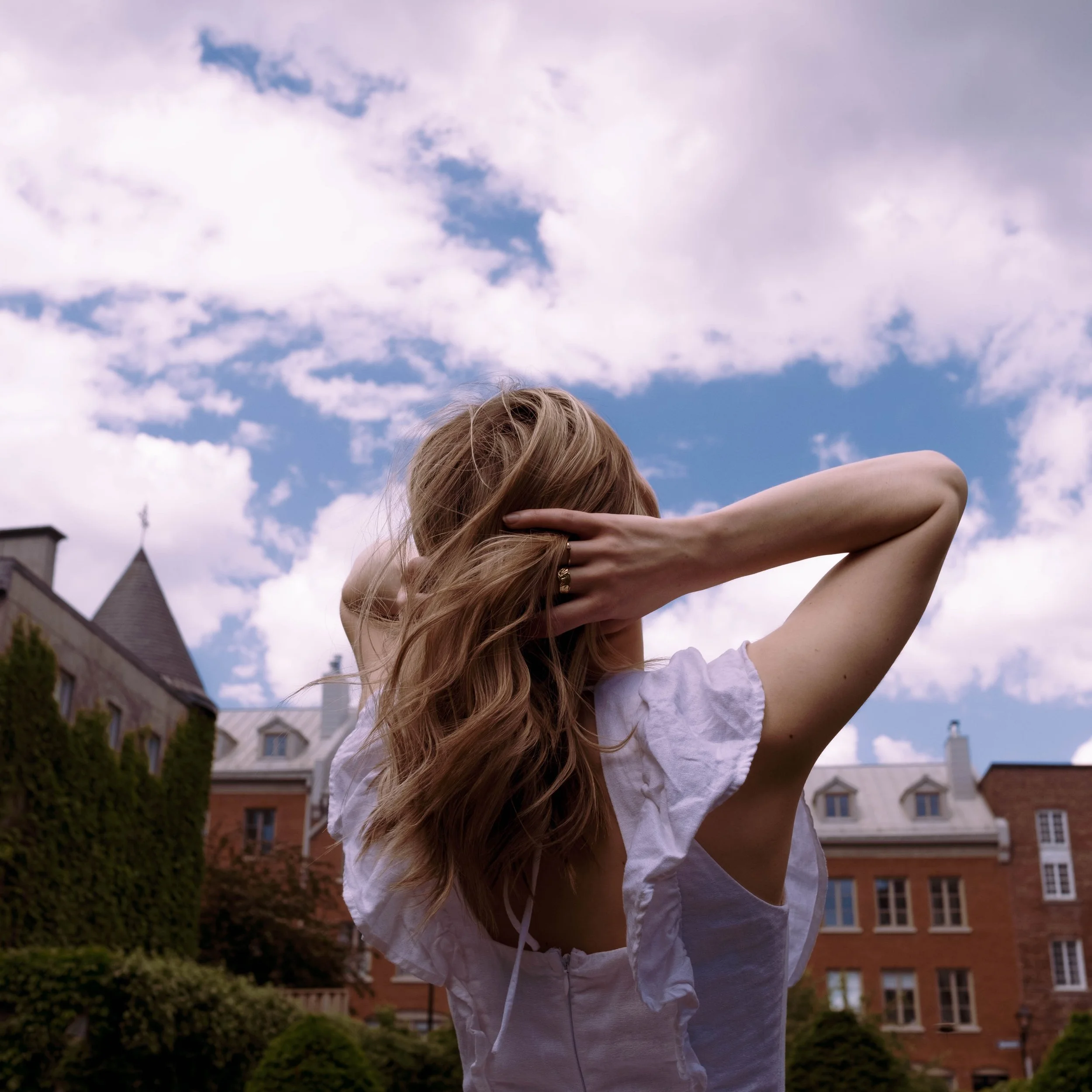 A woman with long, wavy hair is standing outdoors under a partly cloudy sky, with her arms raised and hands touching her hair. She is wearing a light-colored blouse, and there are buildings and greenery in the background.
