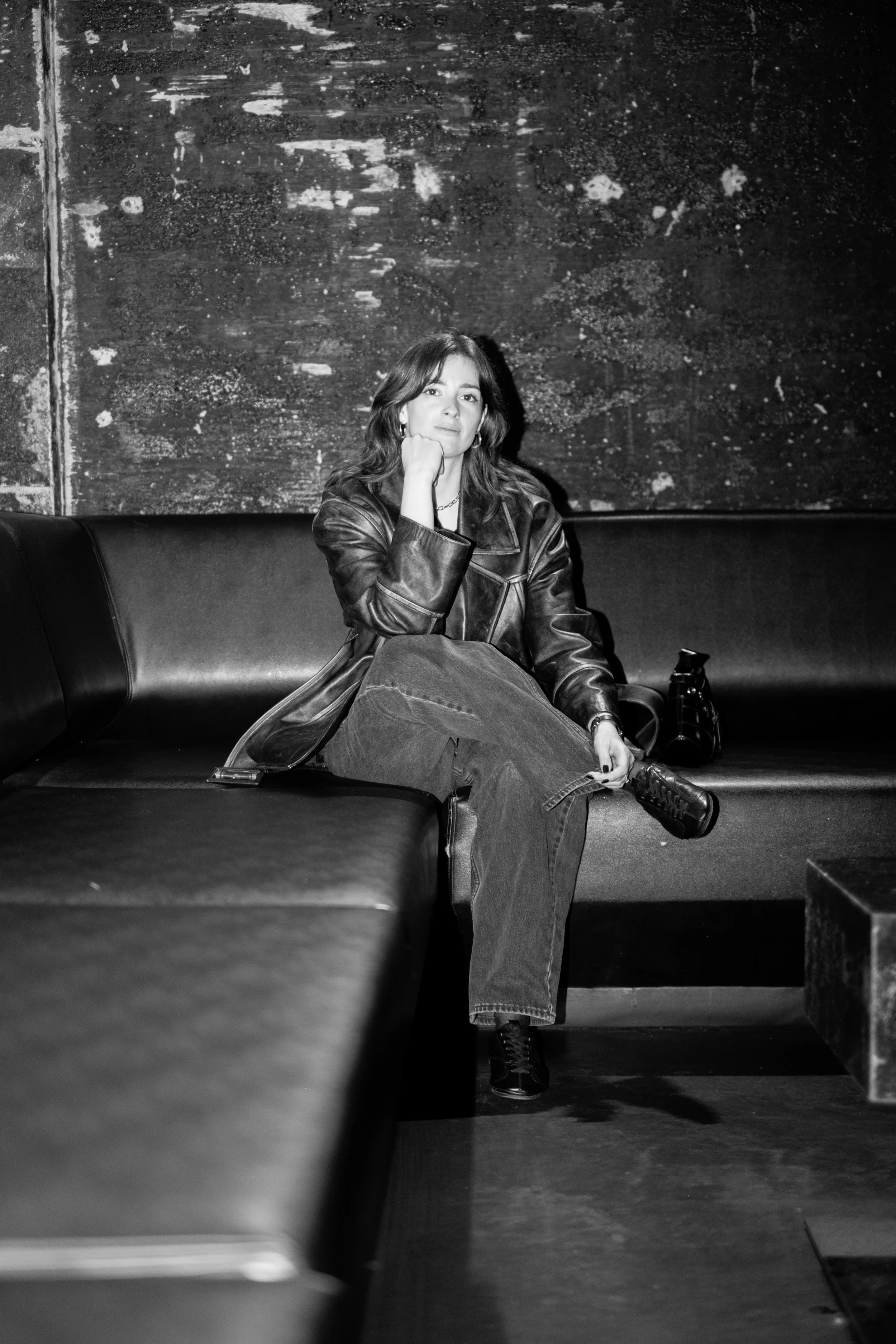 Black and white photo of a woman sitting on a cushioned bench, wearing a leather jacket, with her right hand resting on her face, in an environment with textured walls.