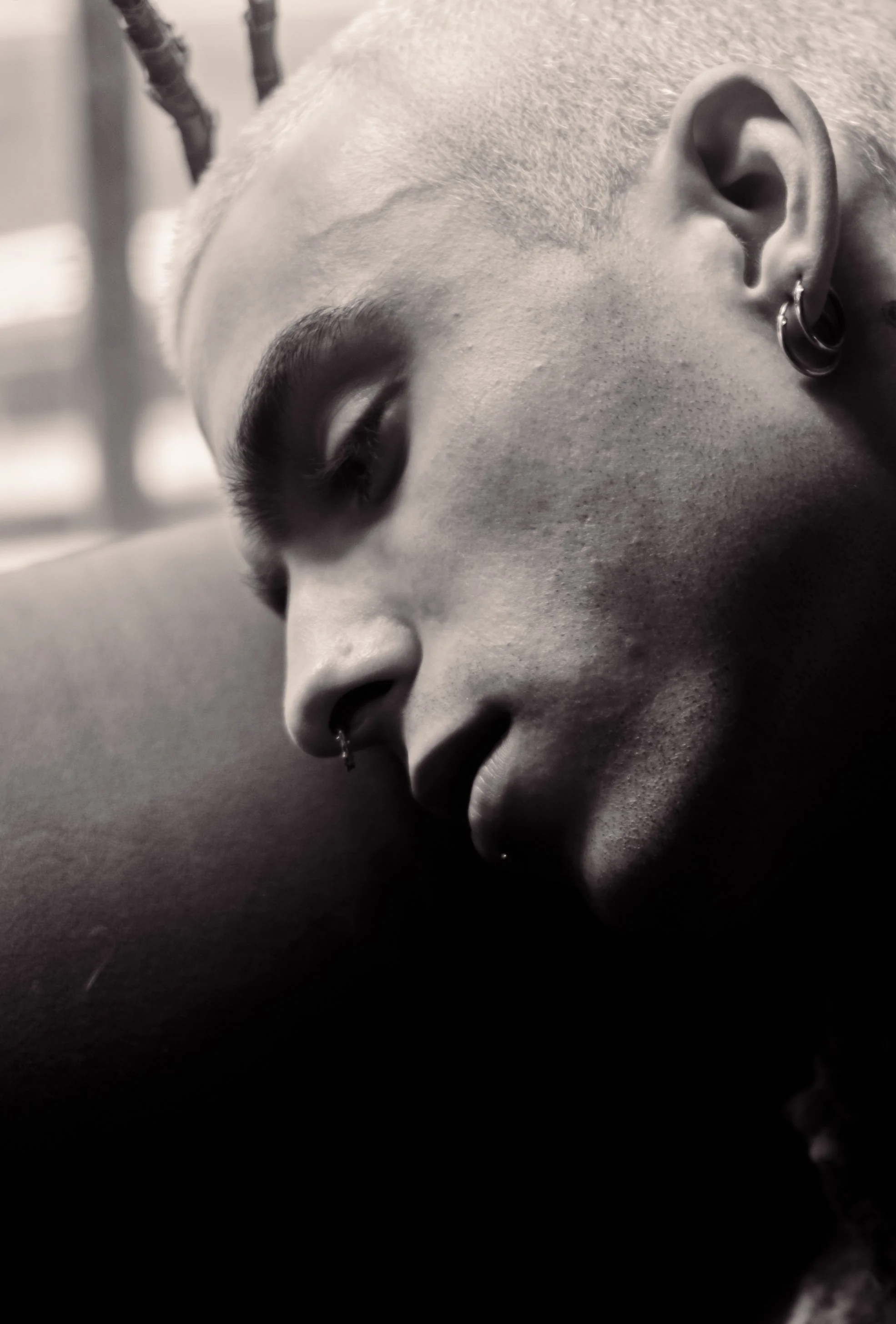 Close-up black and white photograph of a person's face, with features such as closed eyes, earrings, and piercings visible in detail.