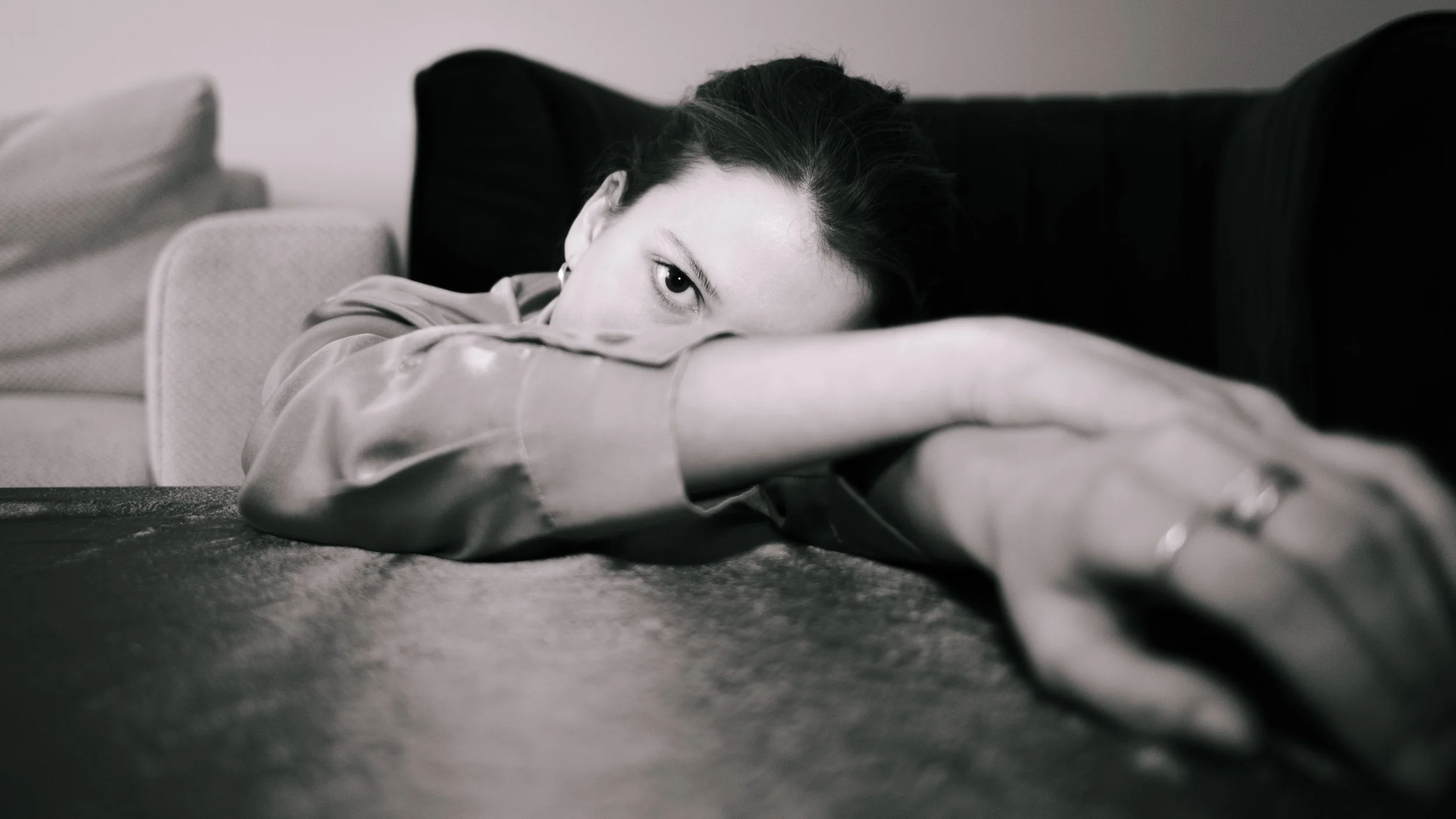 A woman lying down on a table with her head resting on her crossed arms, looking at the camera with one eye visible, in black and white.