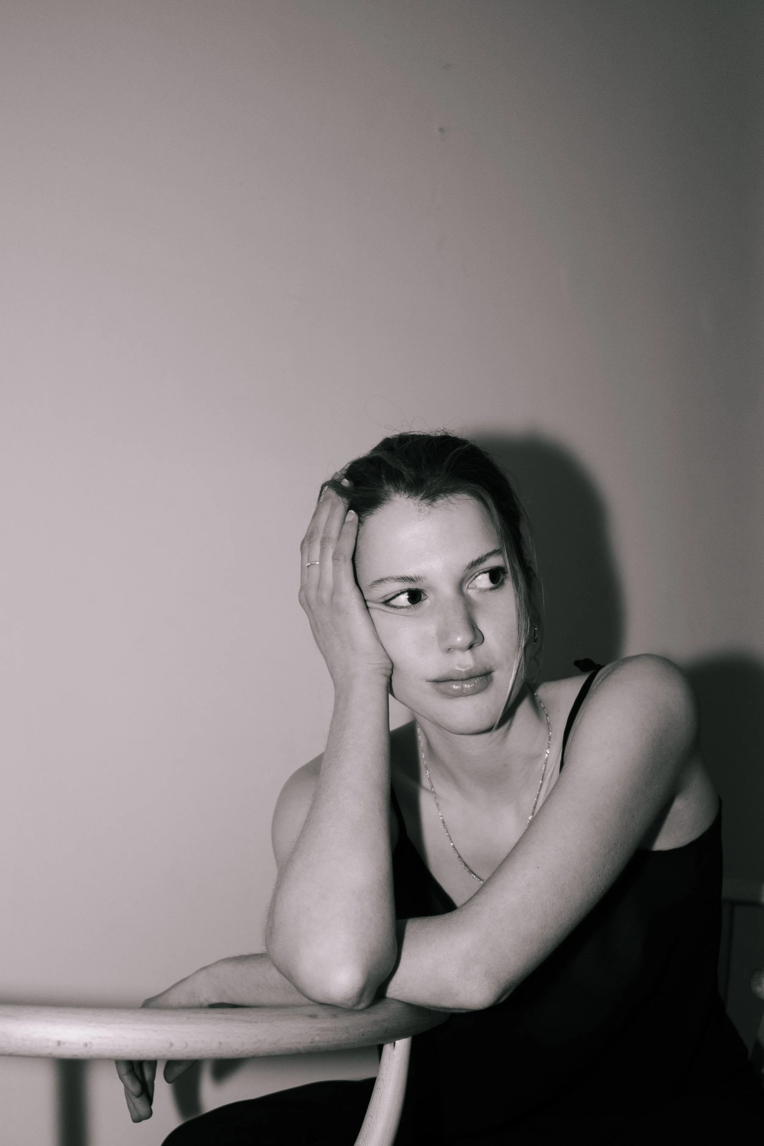 Black and white portrait of a young woman with dark hair, wearing a spaghetti strap top, resting her head on her hand, sitting next to a table.