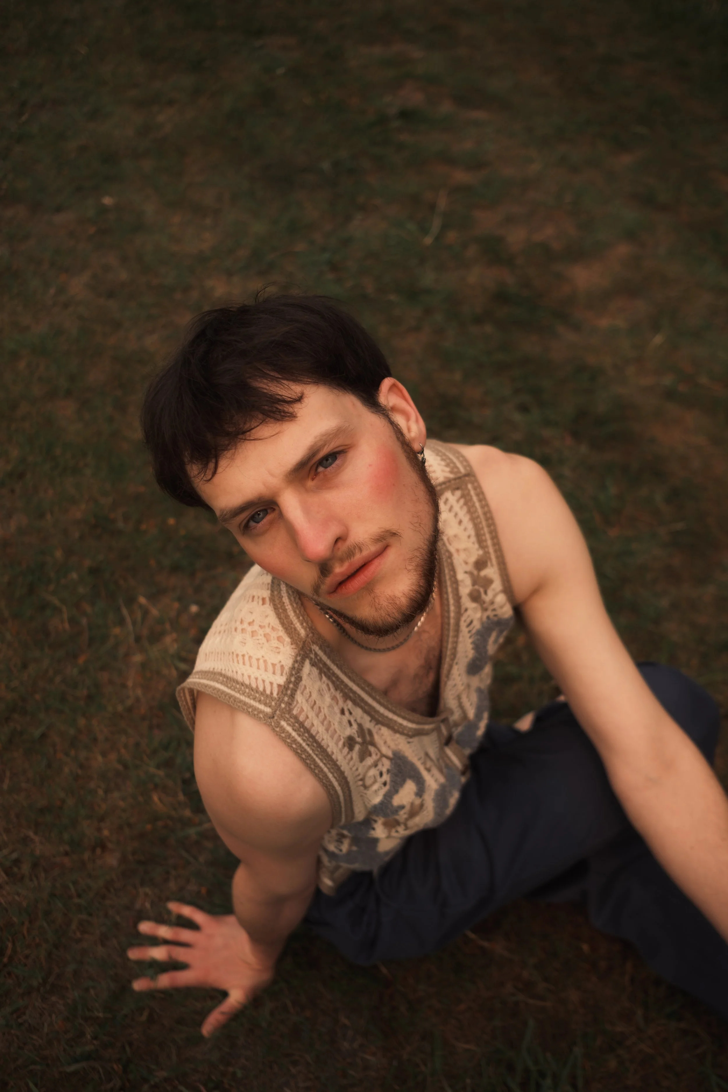 A young man with dark hair and a beard, sitting outdoors on grass. He is looking up at the camera, wearing a sleeveless, patterned shirt. The image is taken from above.