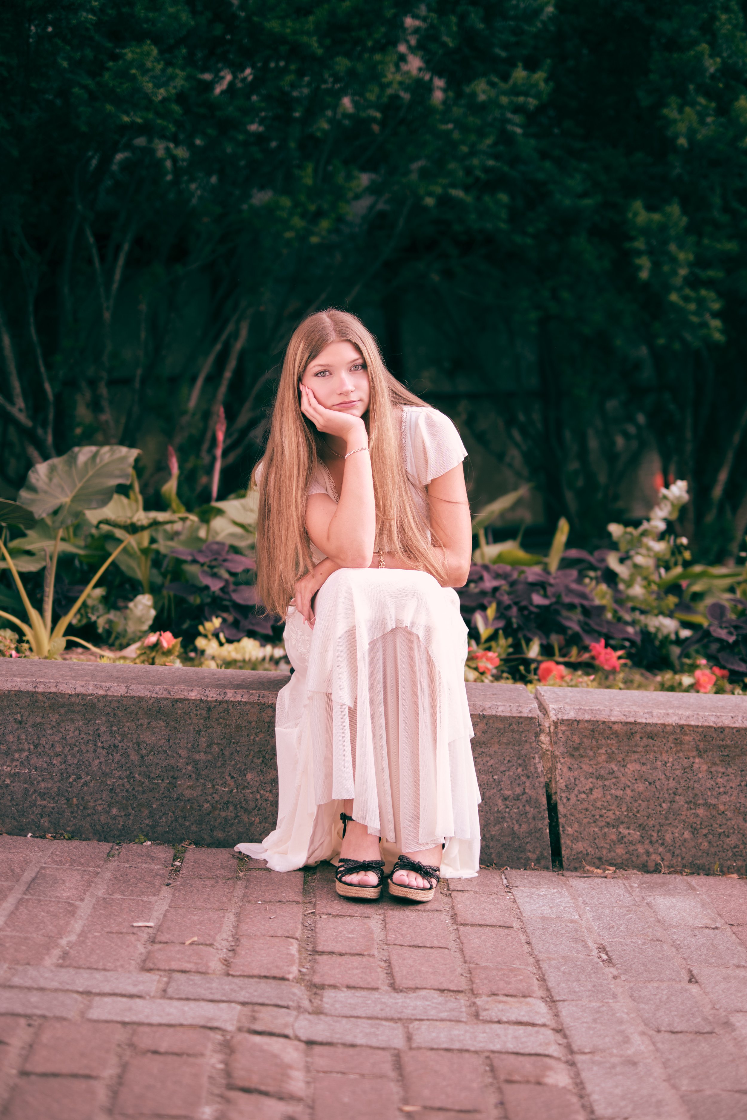 A young woman with long blonde hair wearing a white dress and black sandals sitting on a stone ledge in a park with trees and flowers in the background.