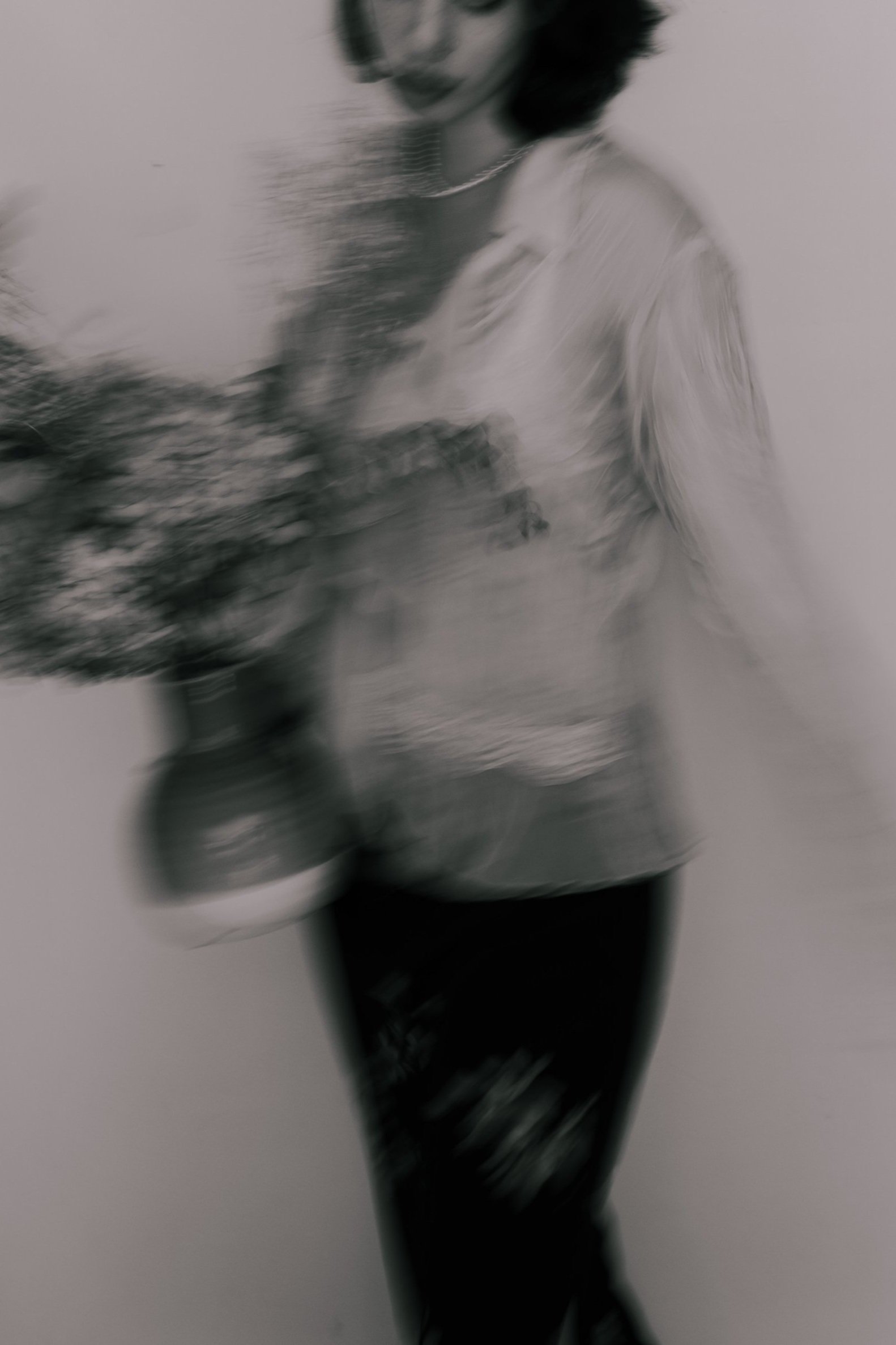A blurred black and white photograph of a woman holding a bouquet of flowers, wearing a striped top and dark pants, with motion blur creating a sense of movement.