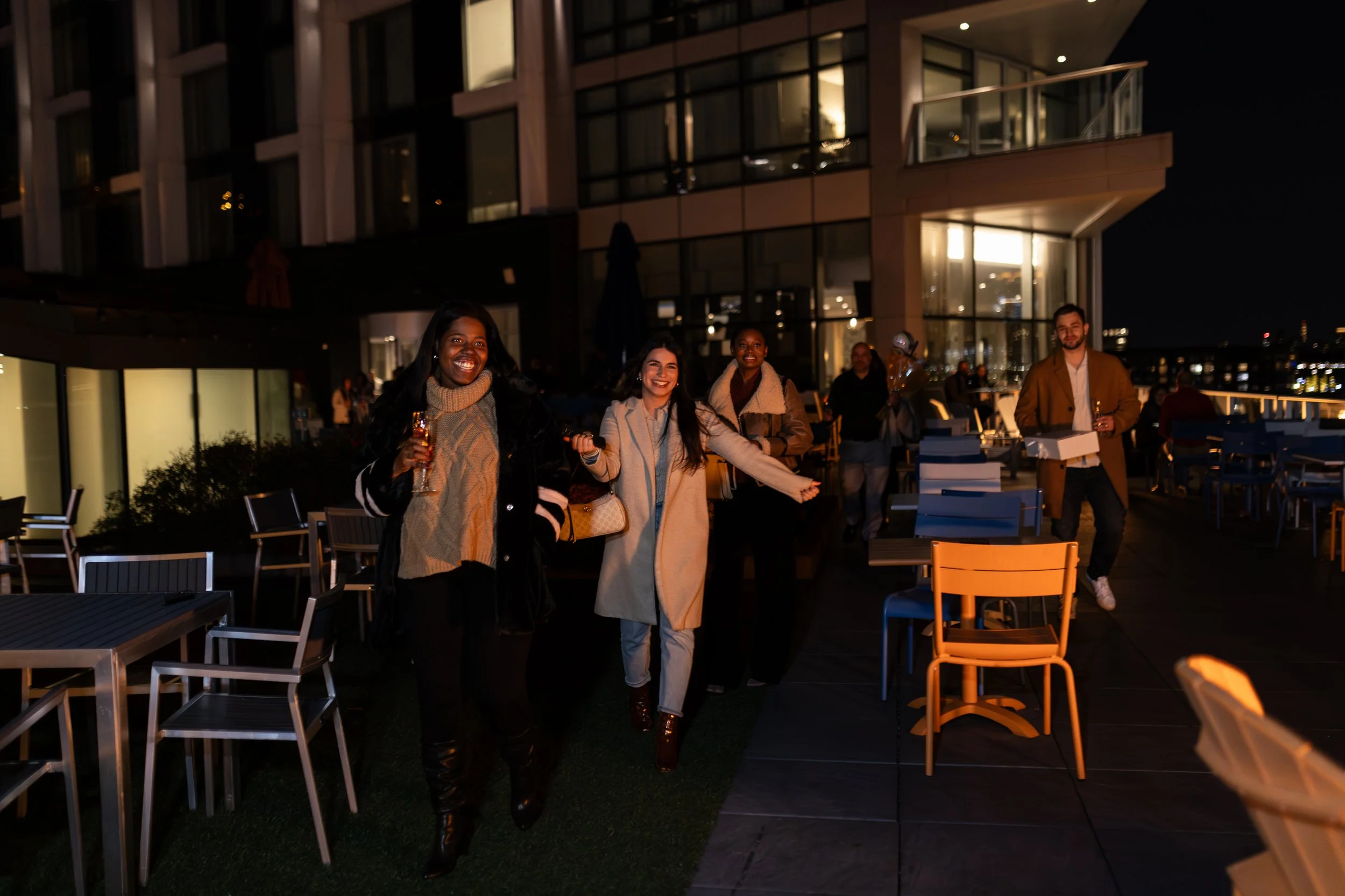 Group of happy people enjoying drinks on a rooftop terrace at night with city lights in the background.
