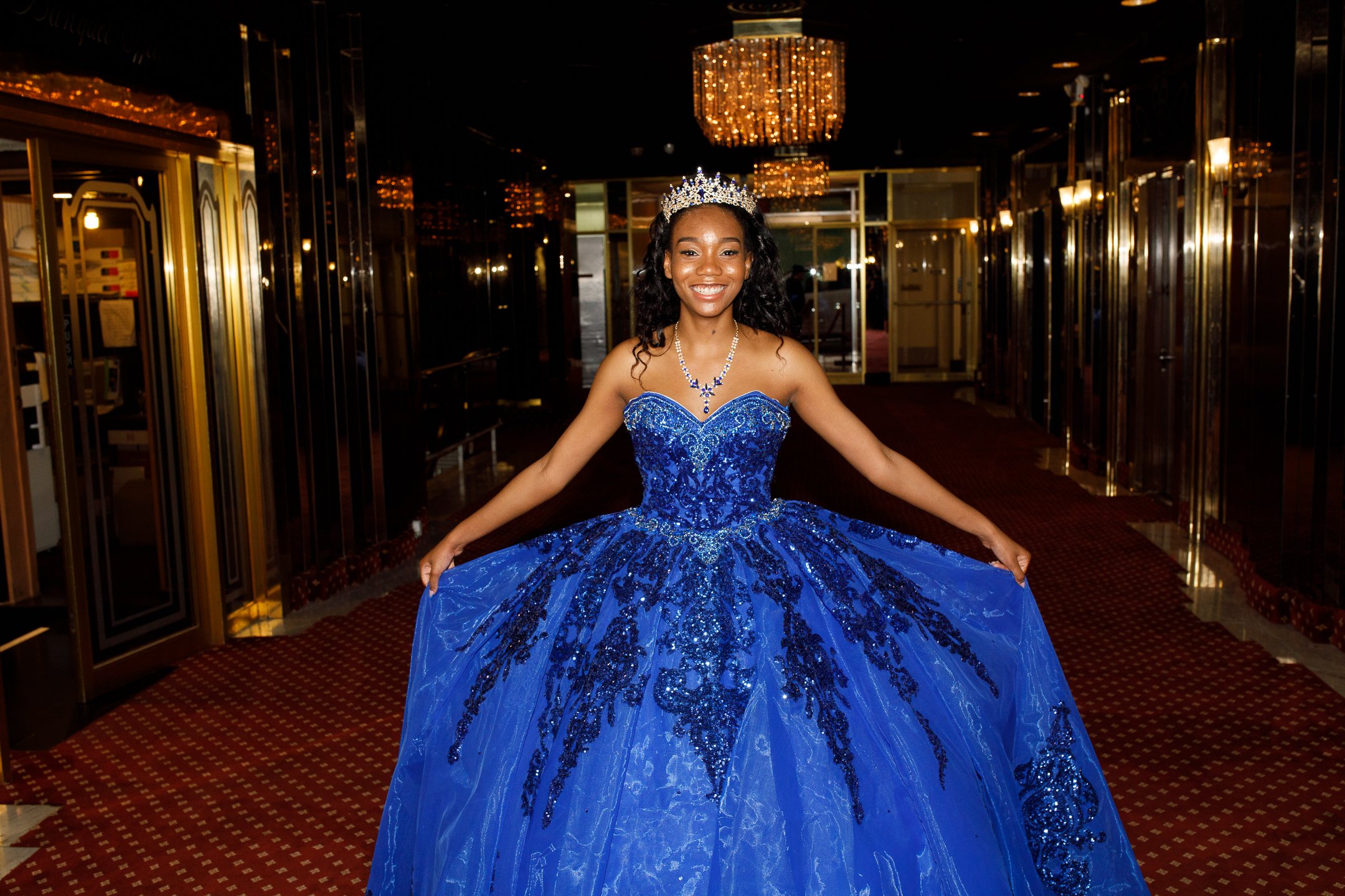 Young woman in a sparkling blue ball gown, wearing a tiara and necklace, smiling in an elegant corridor with reflective walls and a chandelier overhead.