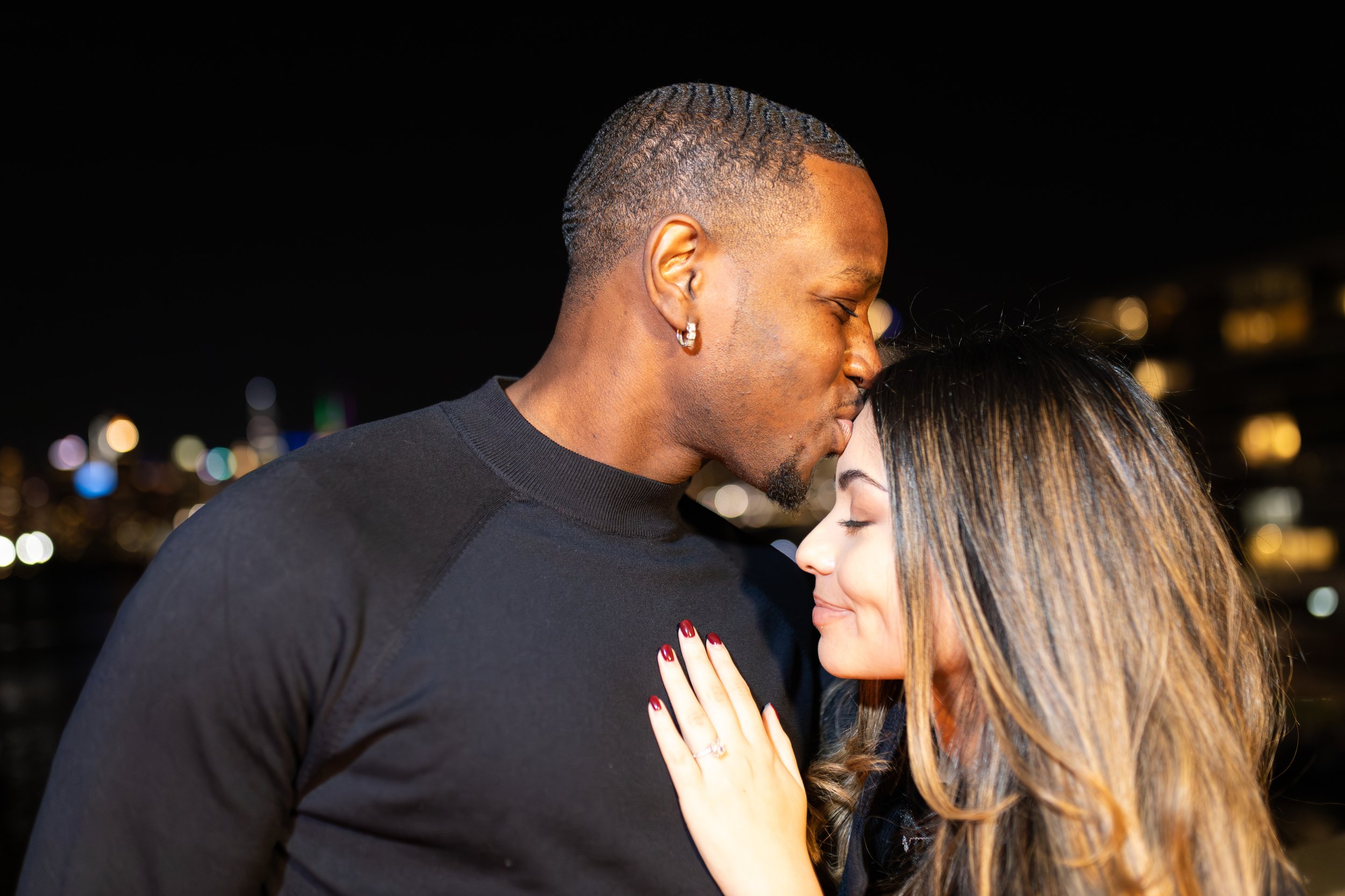 A couple close together at night, with the man gently kissing the woman on her forehead, city lights in the blurred background.