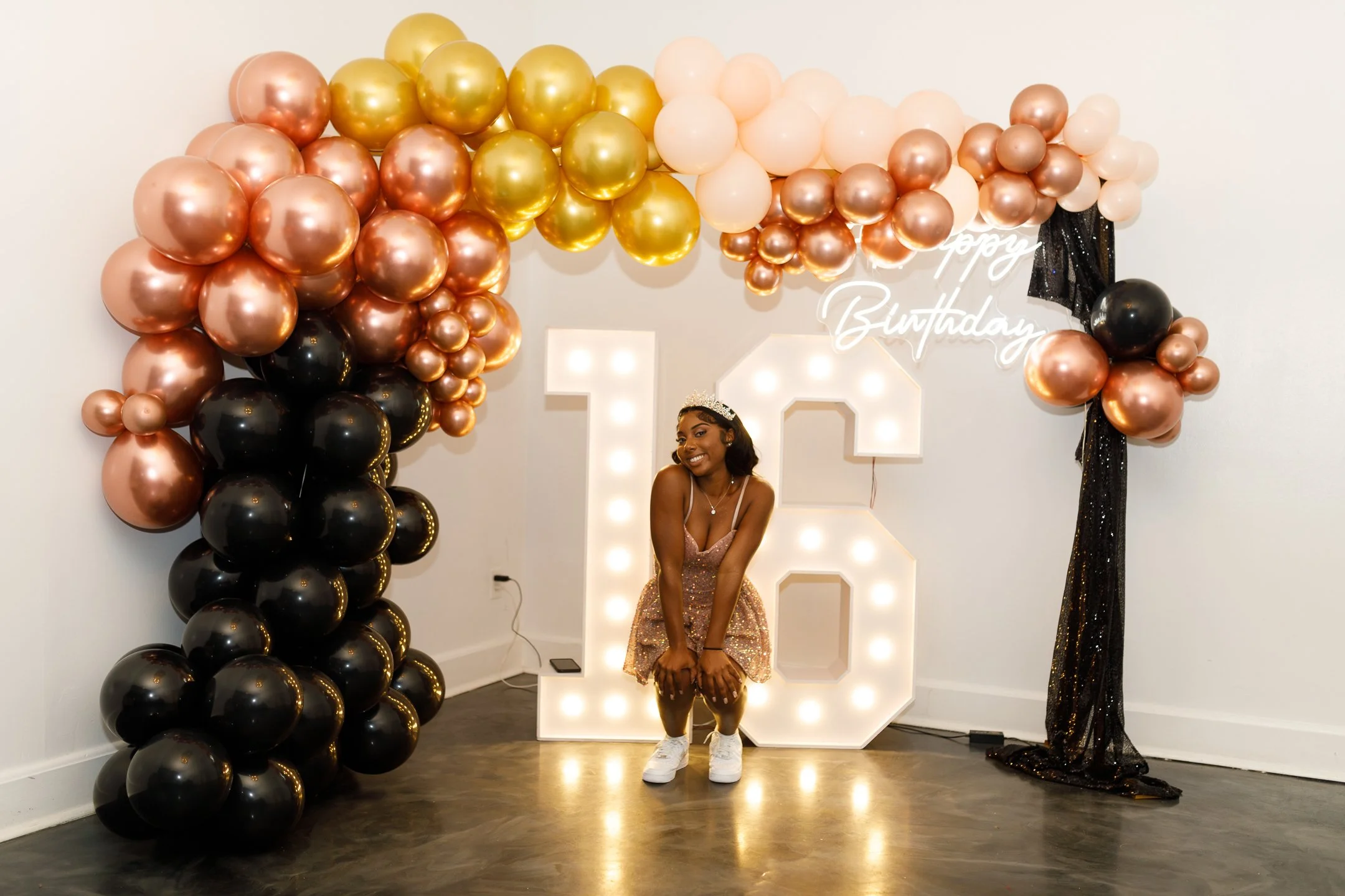 Young woman with a tiara, smiling, at her 16th birthday celebration with a balloon arch in shades of rose gold, gold, black, and pink, and a large illuminated '16' sign, with a 'happy birthday' sign in the background.