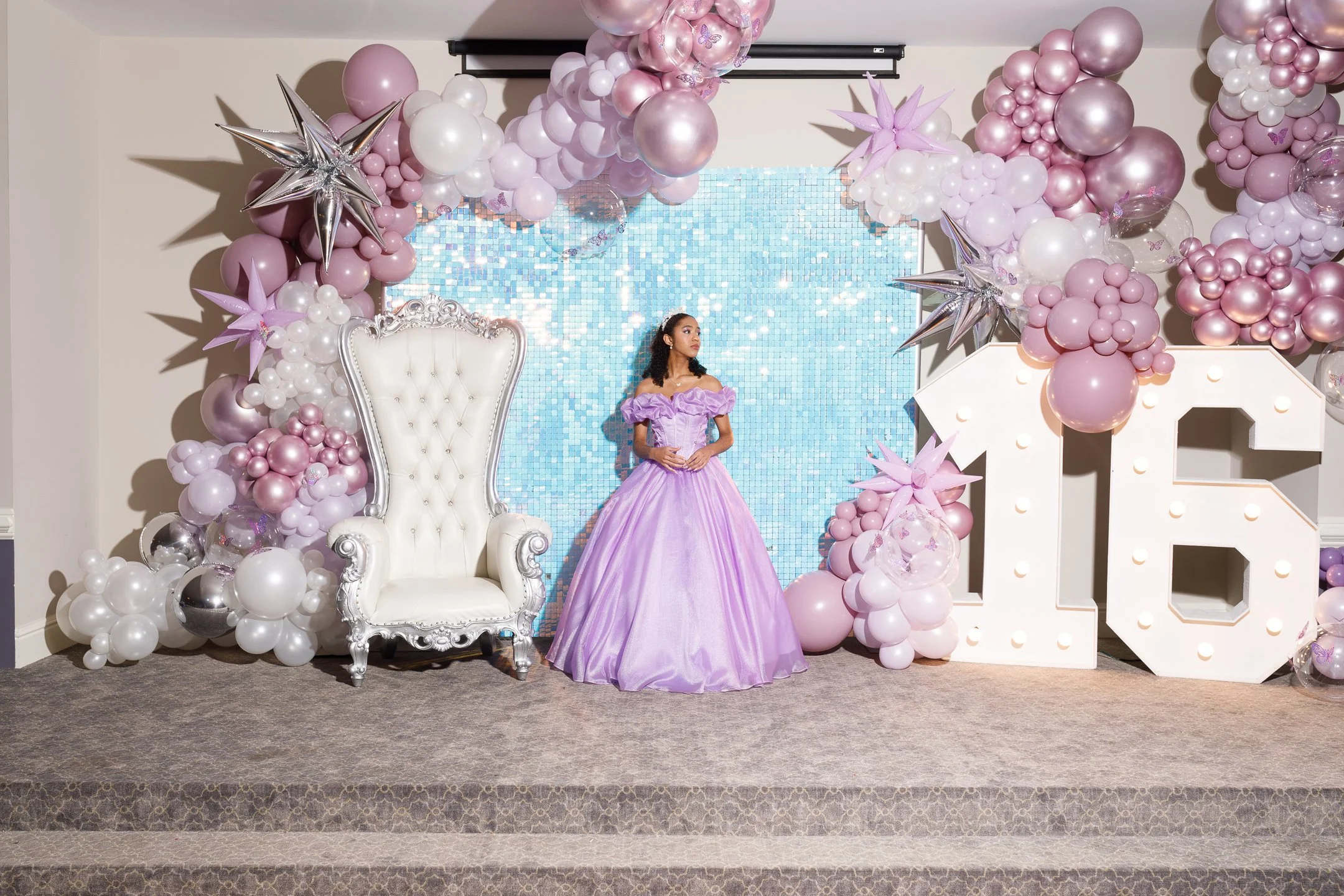 A young woman in a lilac ball gown standing on a decorated stage with pink, white, and silver balloons and a large illuminated number '16' for a sweet 16 birthday celebration.