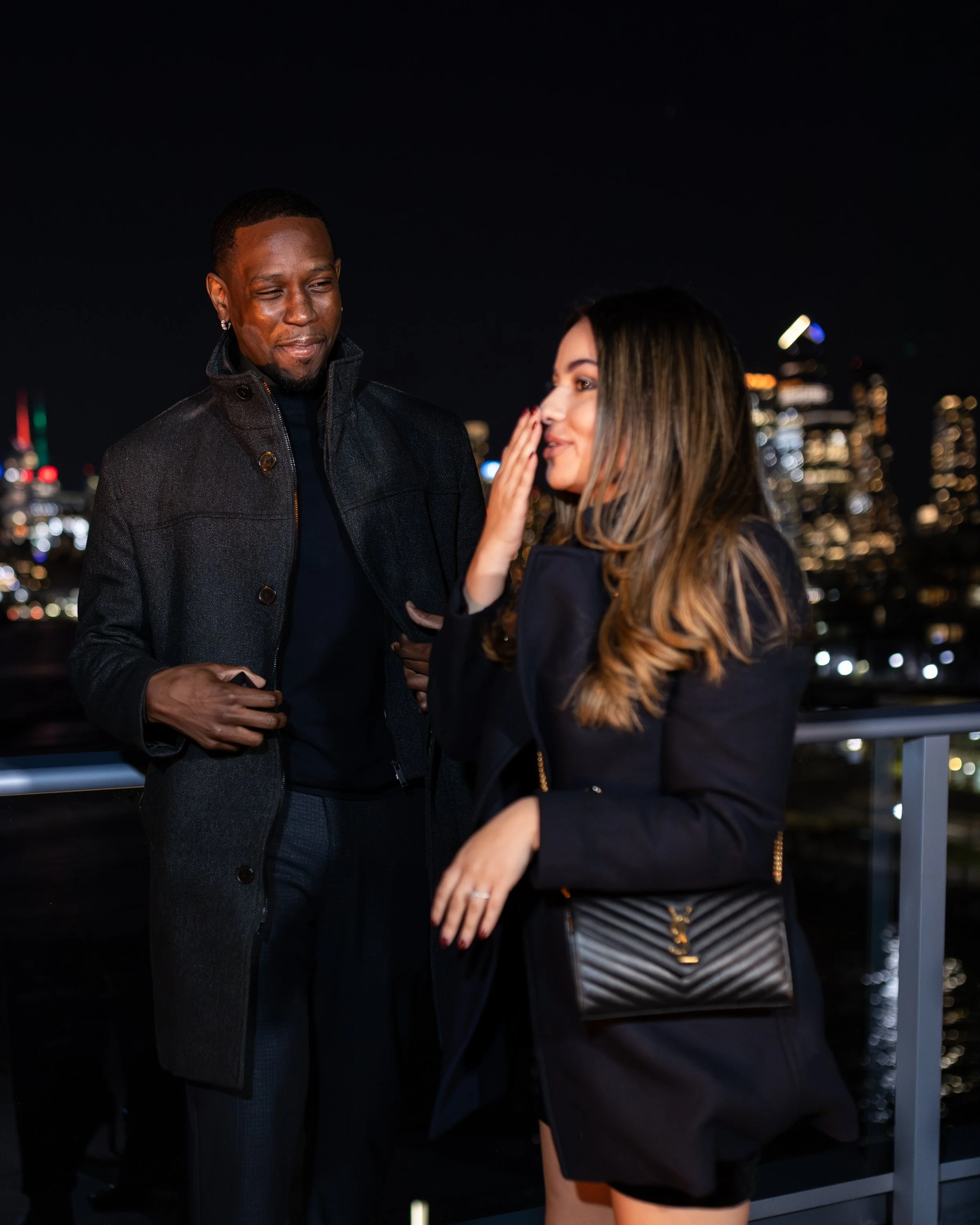 A man and a woman are having a conversation on a rooftop at night, with a city skyline illuminated in the background.