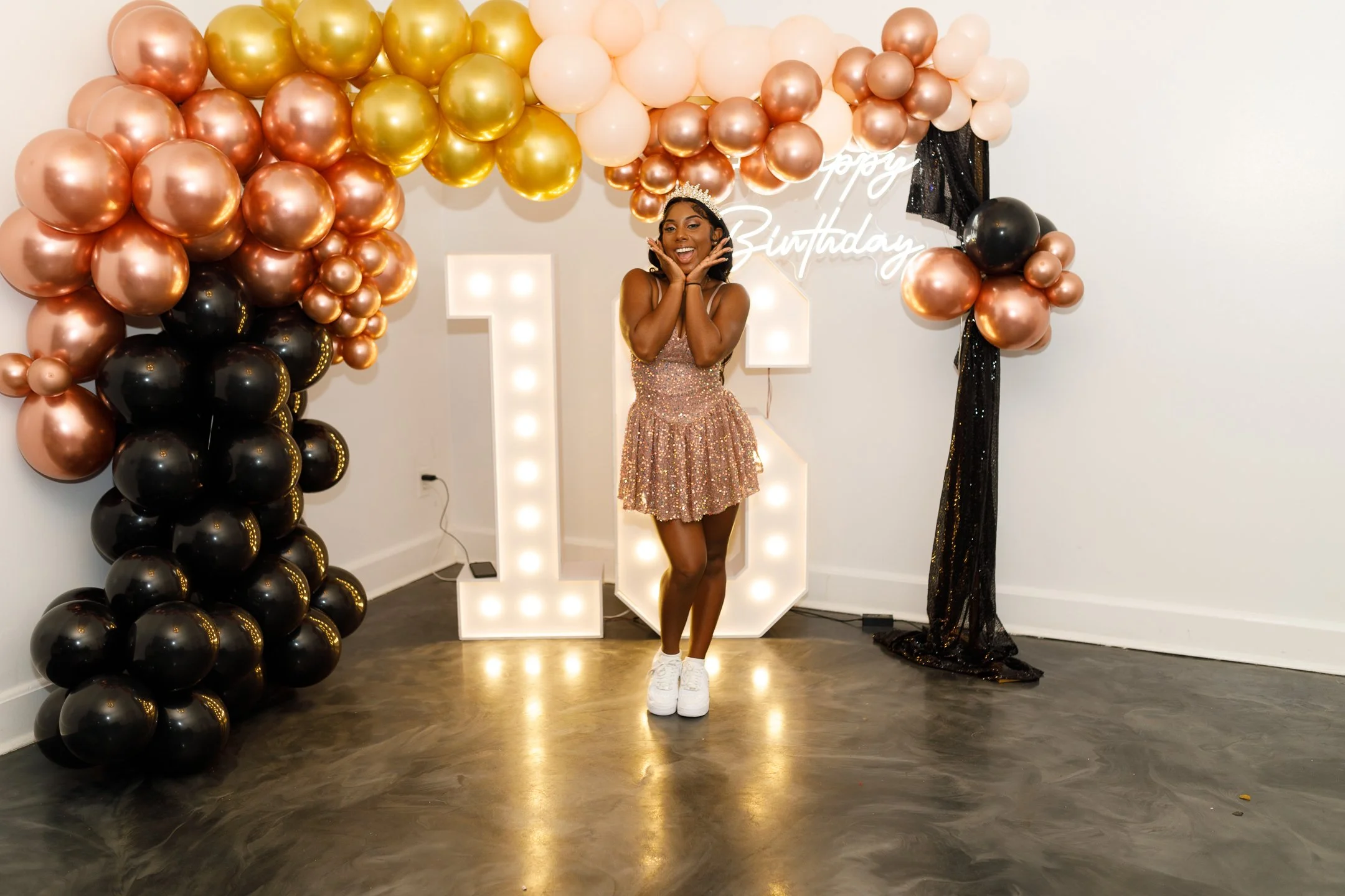 Young woman celebrating birthday standing in front of balloon decorations, large illuminated number '16', and a sign that says 'Happy Birthday'.