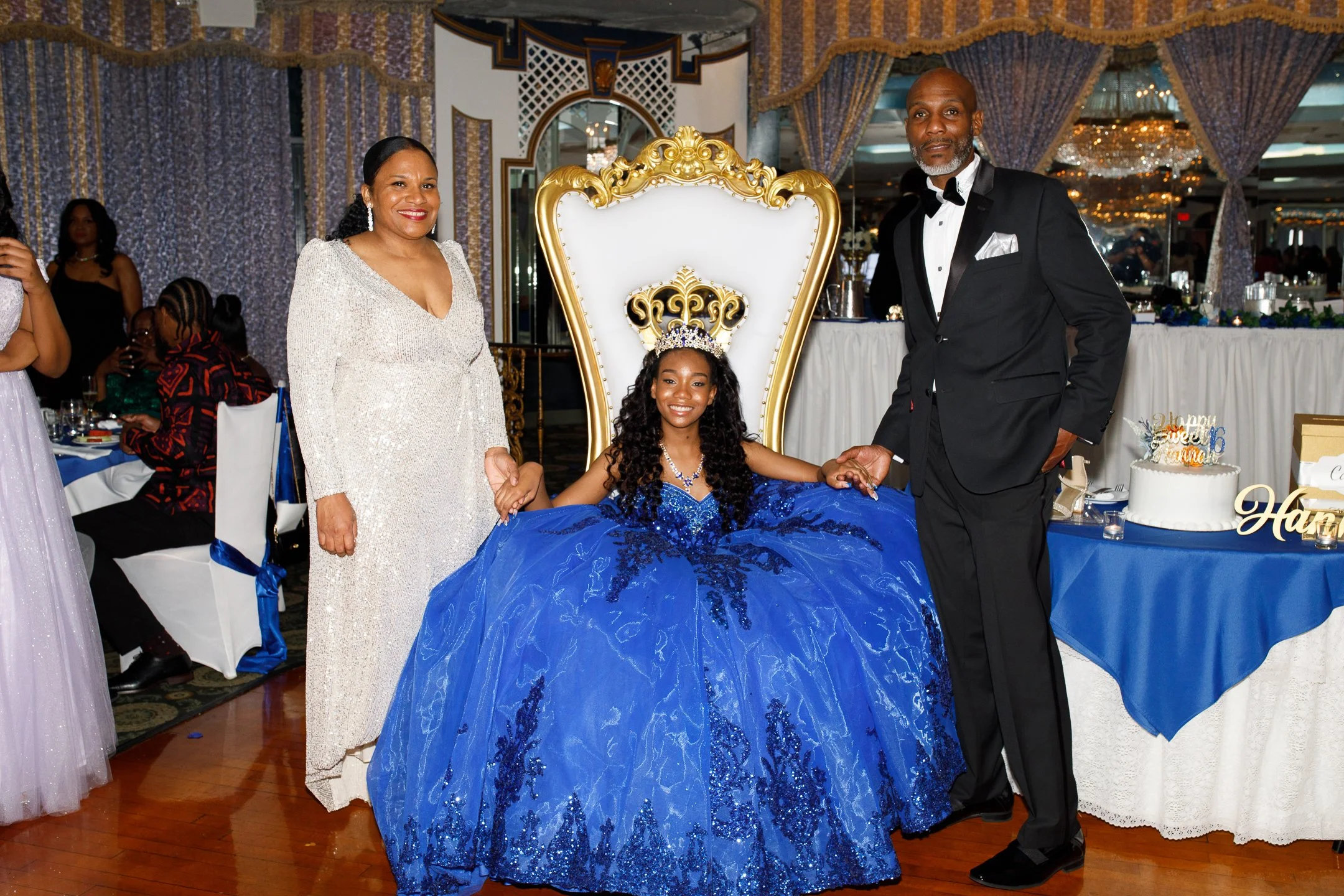 A girl in a blue ball gown sitting on a throne-like chair, flanked by a woman in a white dress and a man in a black tuxedo, at a celebration event with a decorated table and birthday cake.