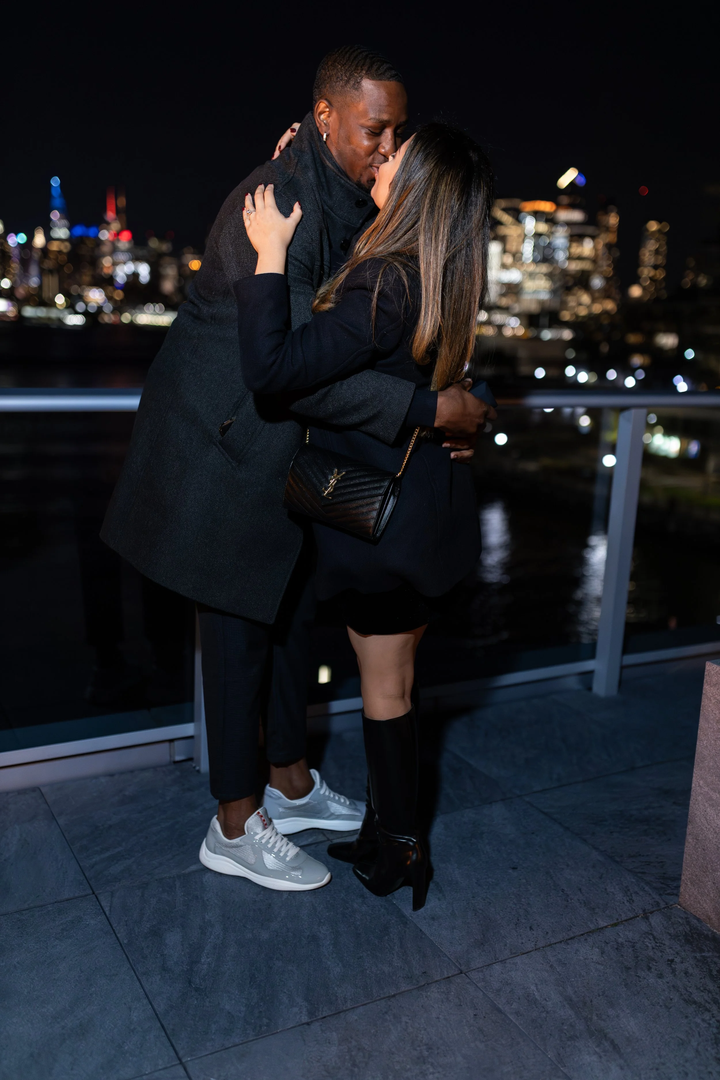 Couple sharing an intimate kiss on a rooftop at night with a city skyline in the background.