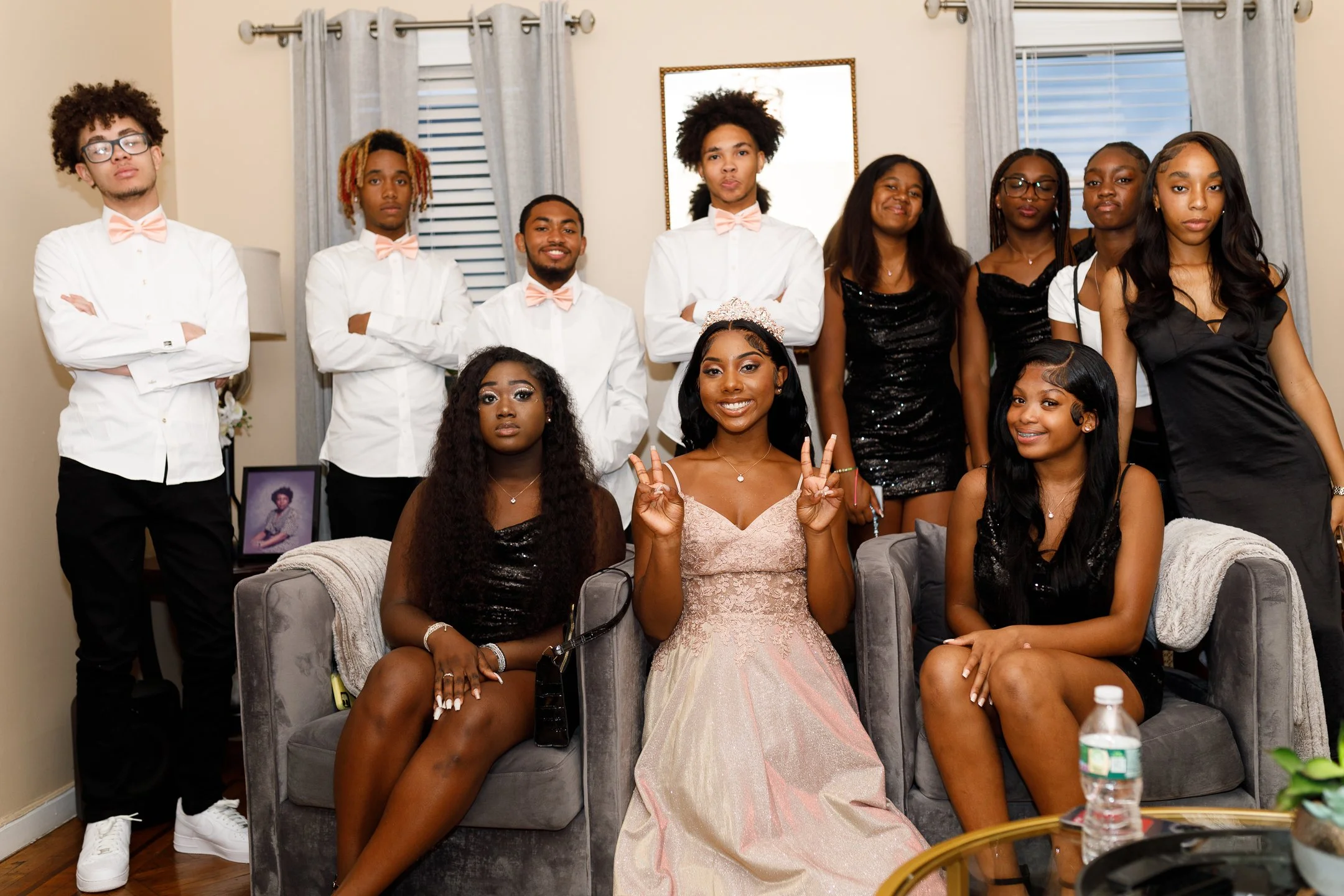 A group of twelve young people, including a woman in a pink dress and tiara, are gathered in a living room, posing for a celebratory photo.