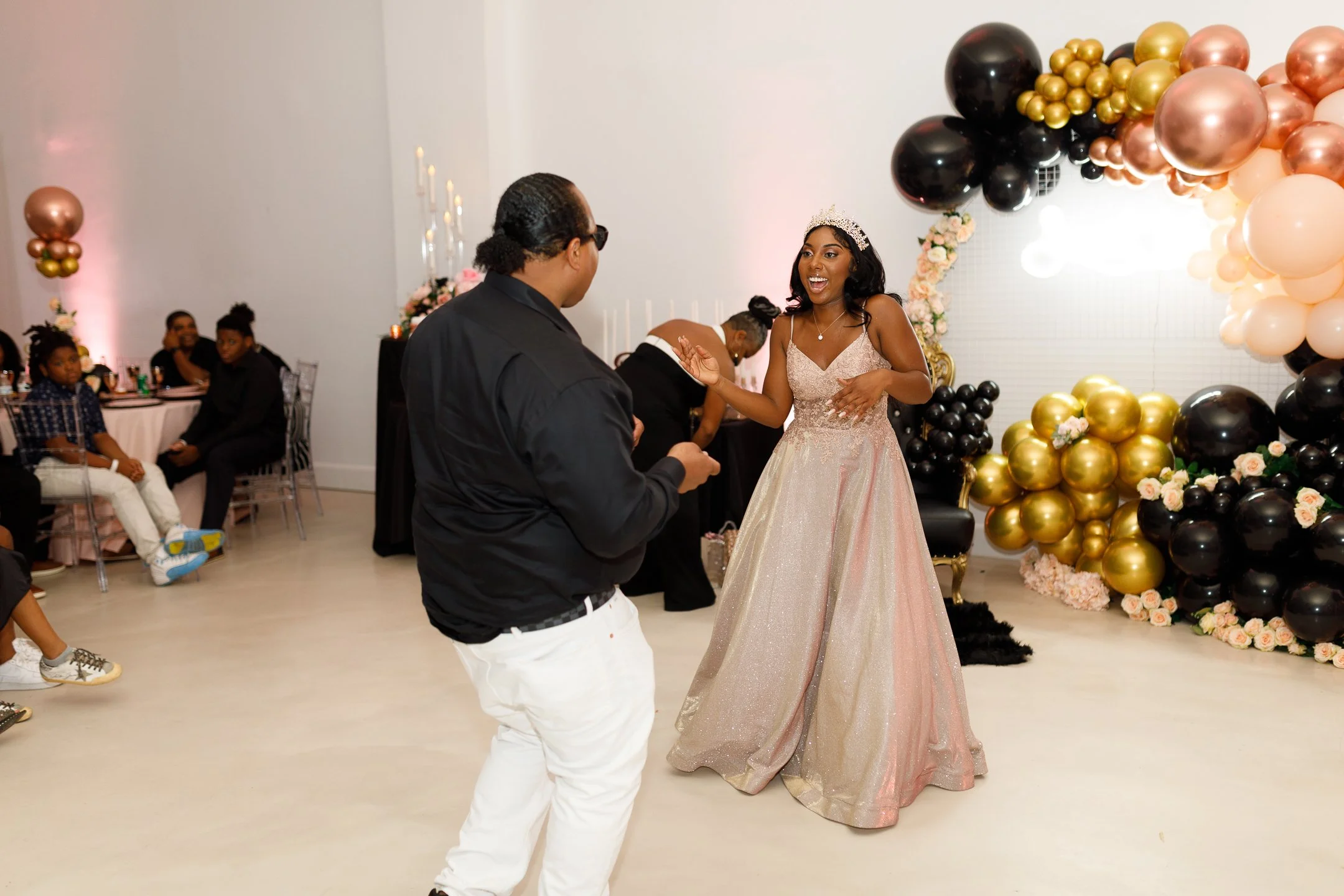 A young woman in a sparkly pink ball gown and tiara is dancing and smiling with a young man in a black shirt and white pants at a celebration event. The background features black, gold, and pink balloon decorations and guests seated at tables.