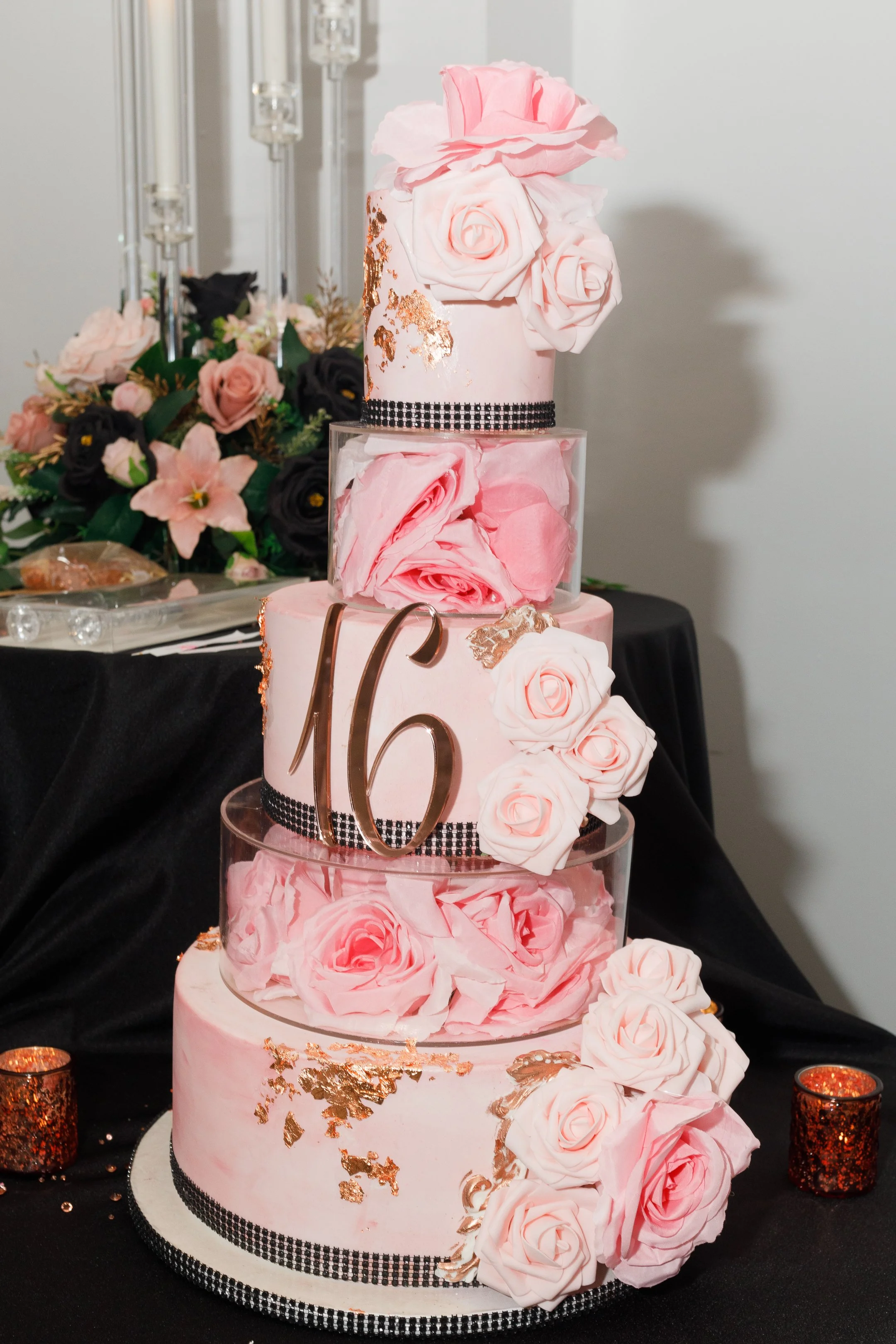 A multi-tiered pink and black wedding cake decorated with pink and white roses, gold accents, and a black and white ribbon, with a large bronze '16' topper on the middle tier.