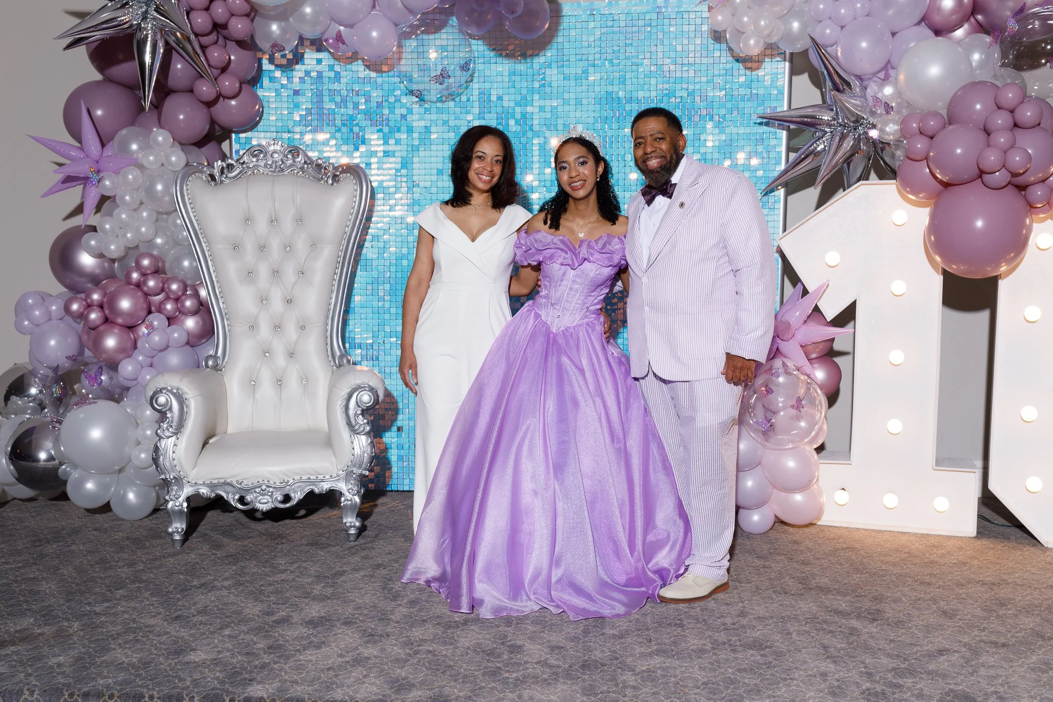A woman in a lavender ball gown standing between a man in a white striped suit and a woman in a white dress, at a celebration with pastel balloon decorations and a blue mosaic backdrop.