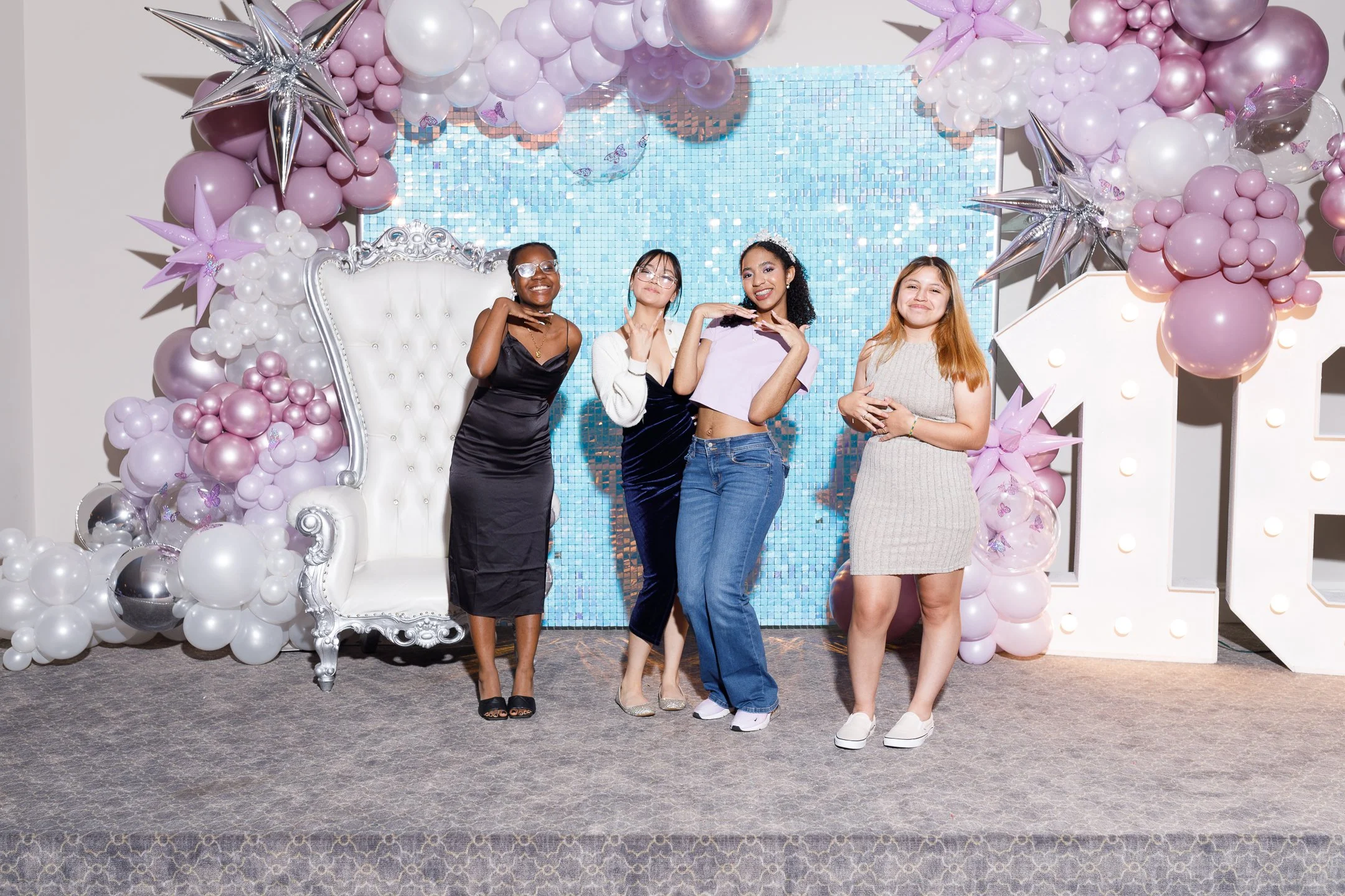 Four young women standing in front of a balloon and light display backdrop at a celebration or party, smiling and posing for the camera.