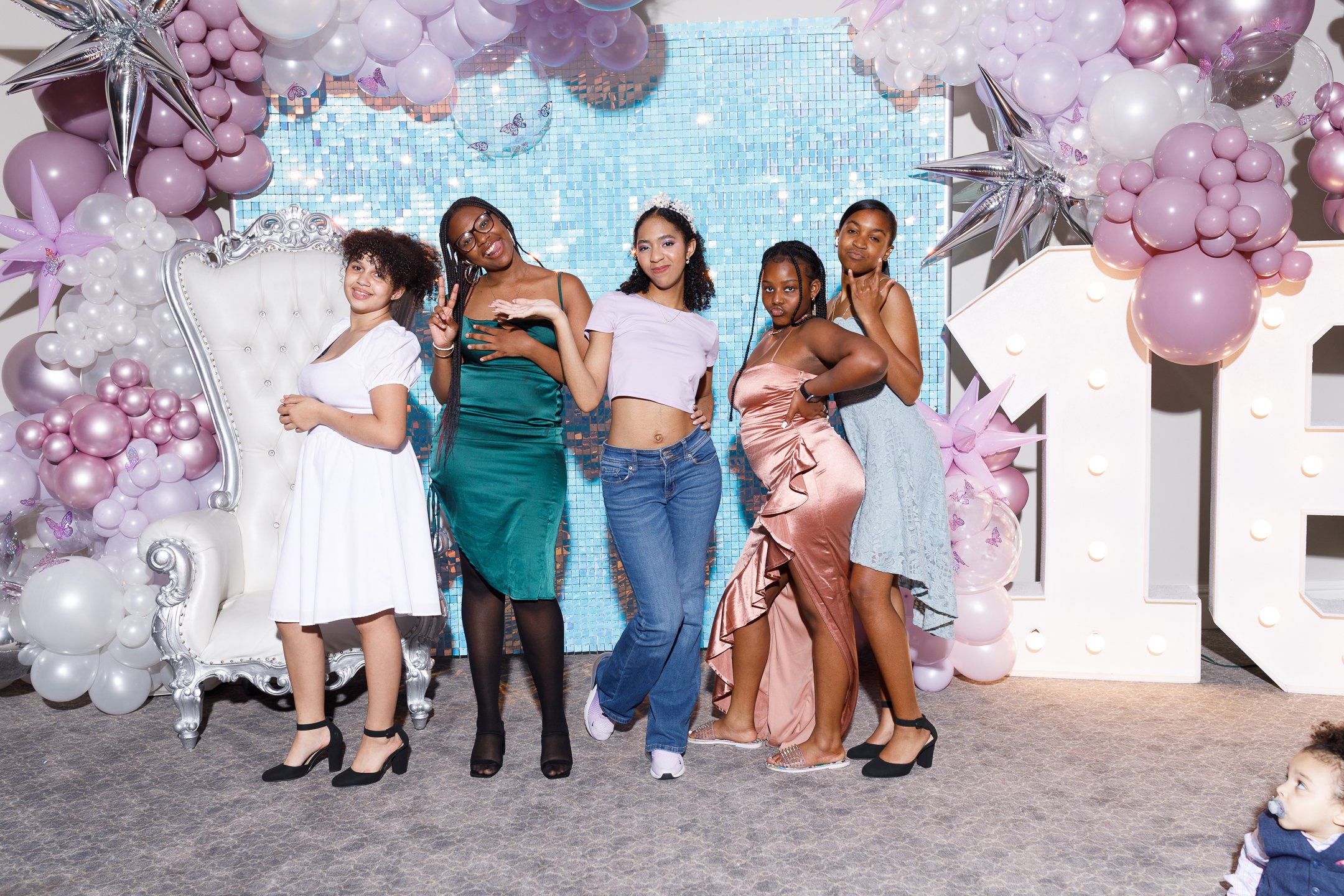Five young women are standing in front of a decorated backdrop with pink and white balloons, stars, and butterfly designs. They are posing and smiling at a celebration or party.