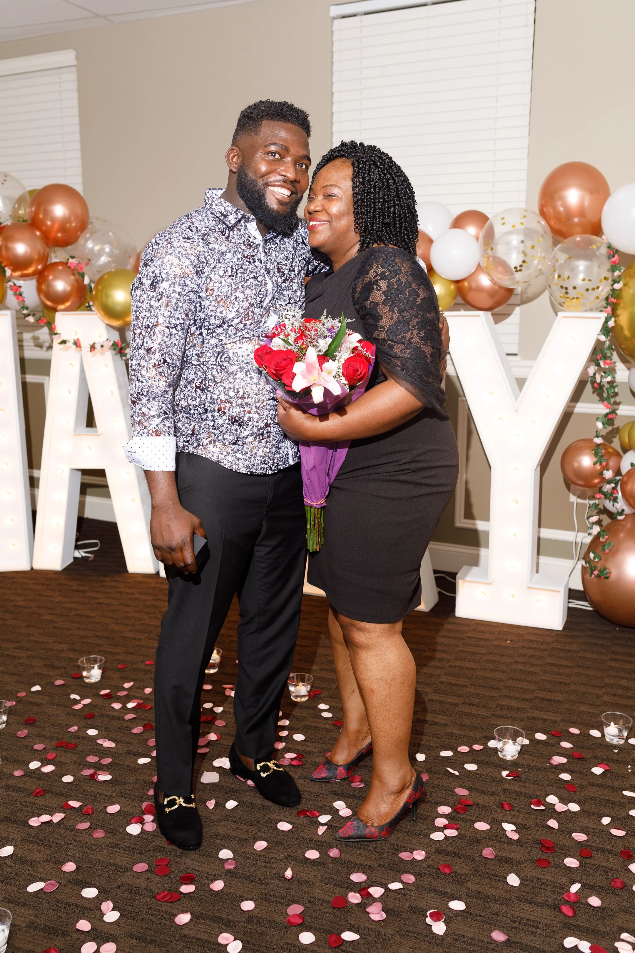 A couple standing together in a decorated room with balloons and large illuminated letters, holding a bouquet of flowers, celebrating a special occasion.