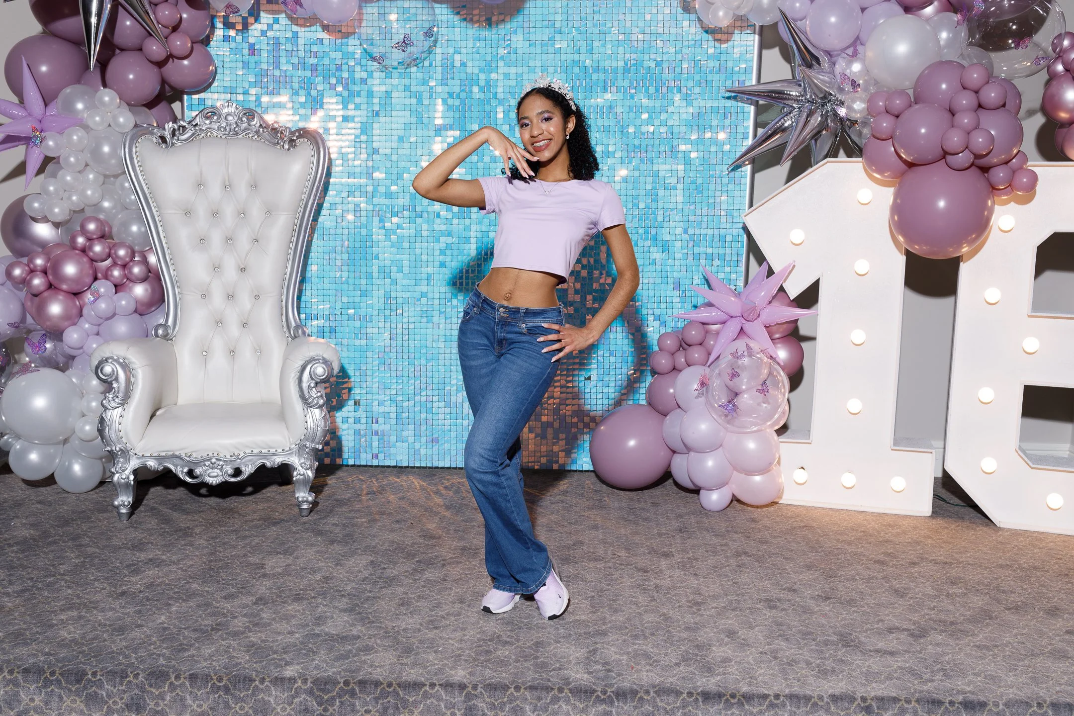 A young woman posing and smiling at a decorated party setup with balloon arrangements, a large white illuminated letter, and a blue tiled wall background.