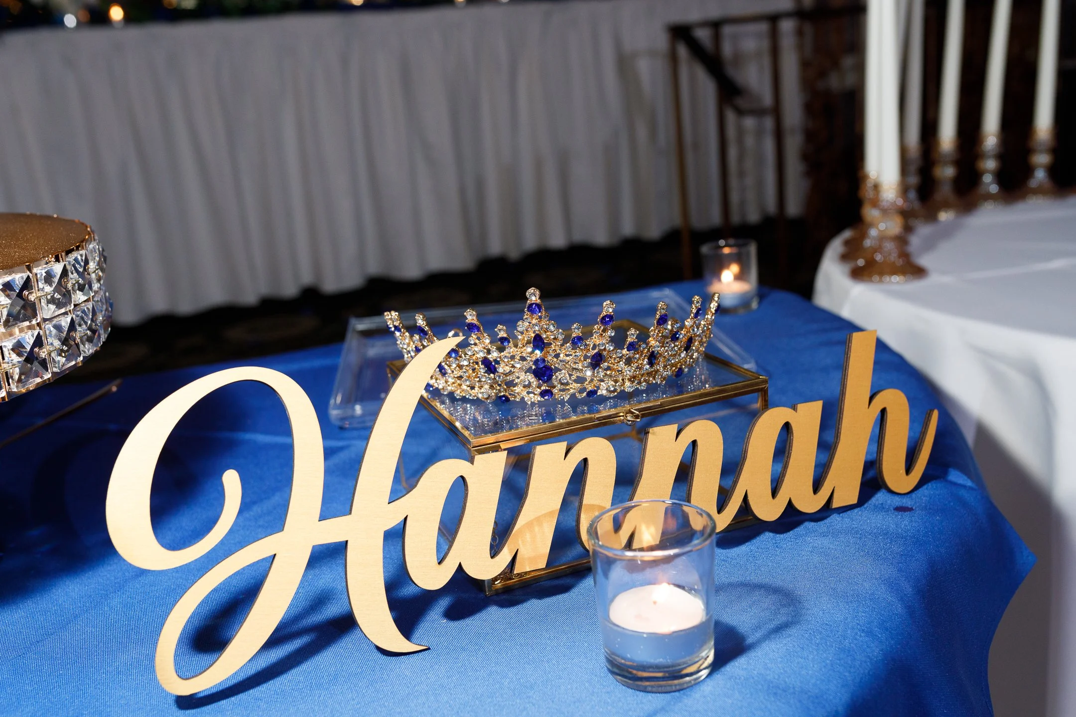Table decorated with a blue tablecloth, a gold sign spelling 'Hannah,' a sparkling crown with blue accents, and several lit candles.