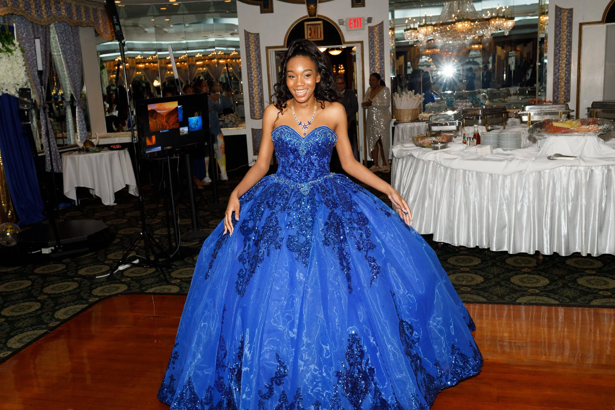 Young woman in a royal blue formal gown with lace and beading, smiling at a celebration or prom event in an elegantly decorated venue.