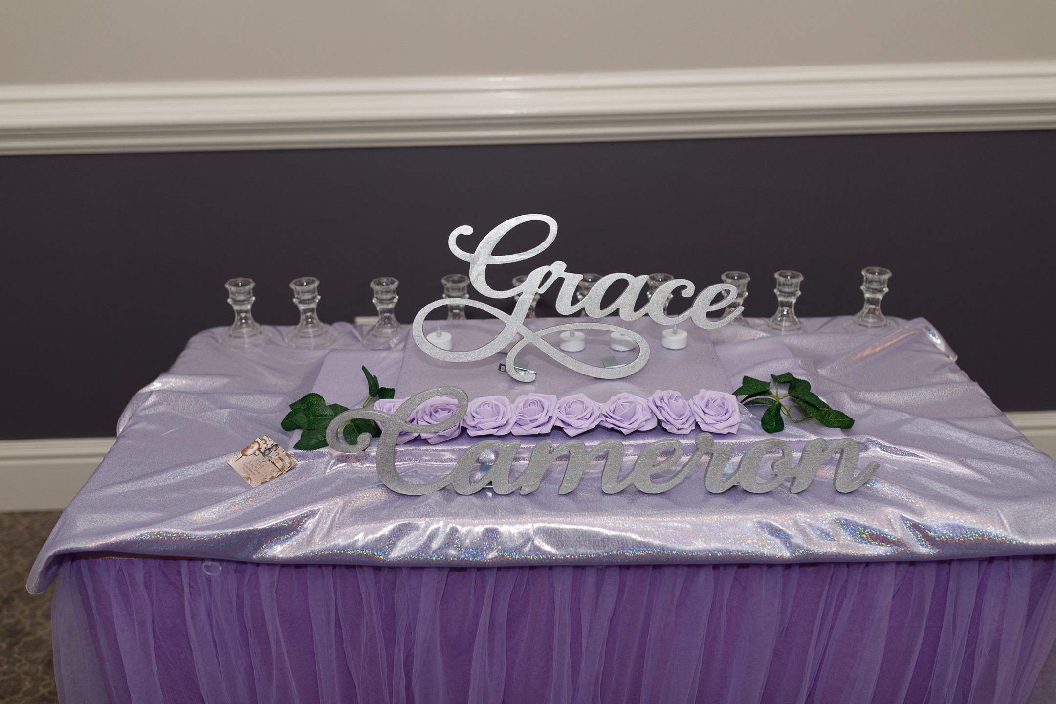 A decorated table with a purple tulle tablecloth, a shiny fabric overlay, and a purple rose arrangement. There is a sign that reads 'Grace & Cameron' and several small bottles behind it.