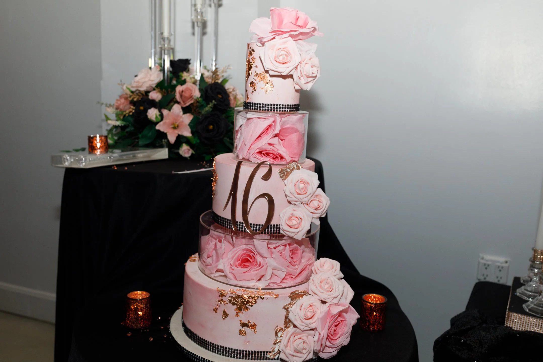 Pink four-tiered wedding cake decorated with pink roses, gold accents, and black ribbon, with a large gold '16' in front, on a black table with candles and floral arrangements in the background.