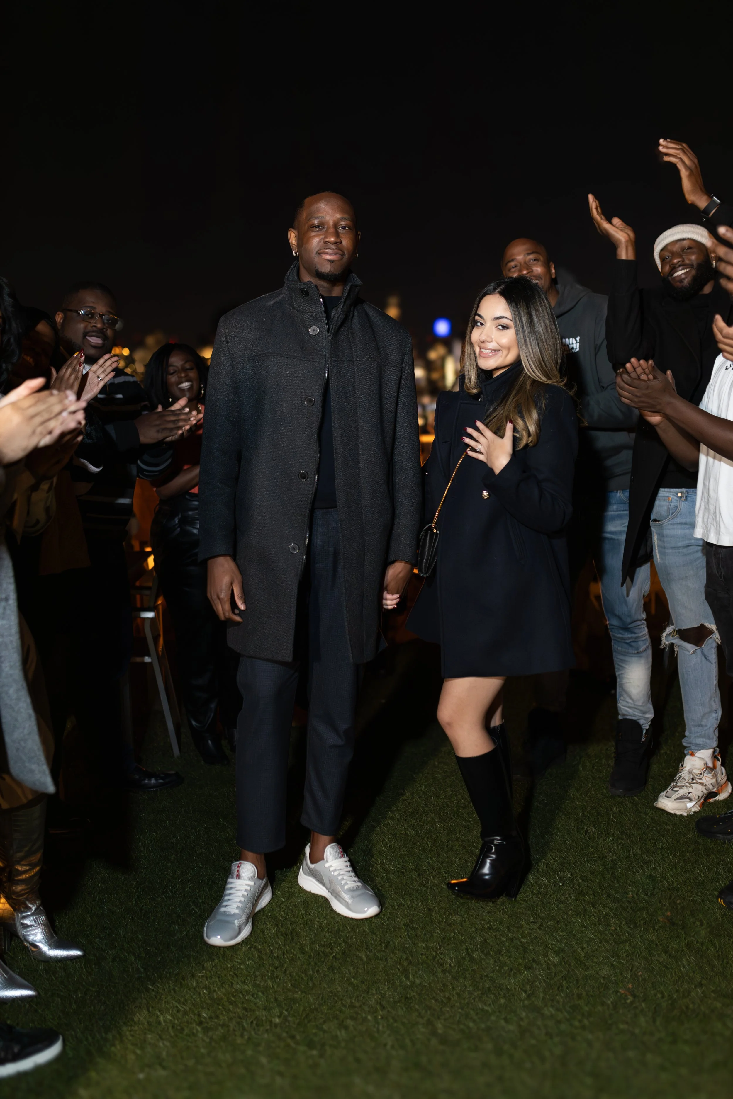 A diverse group of people celebrating outdoors at night, with two individuals in the foreground holding hands, surrounded by clapping and smiling friends.