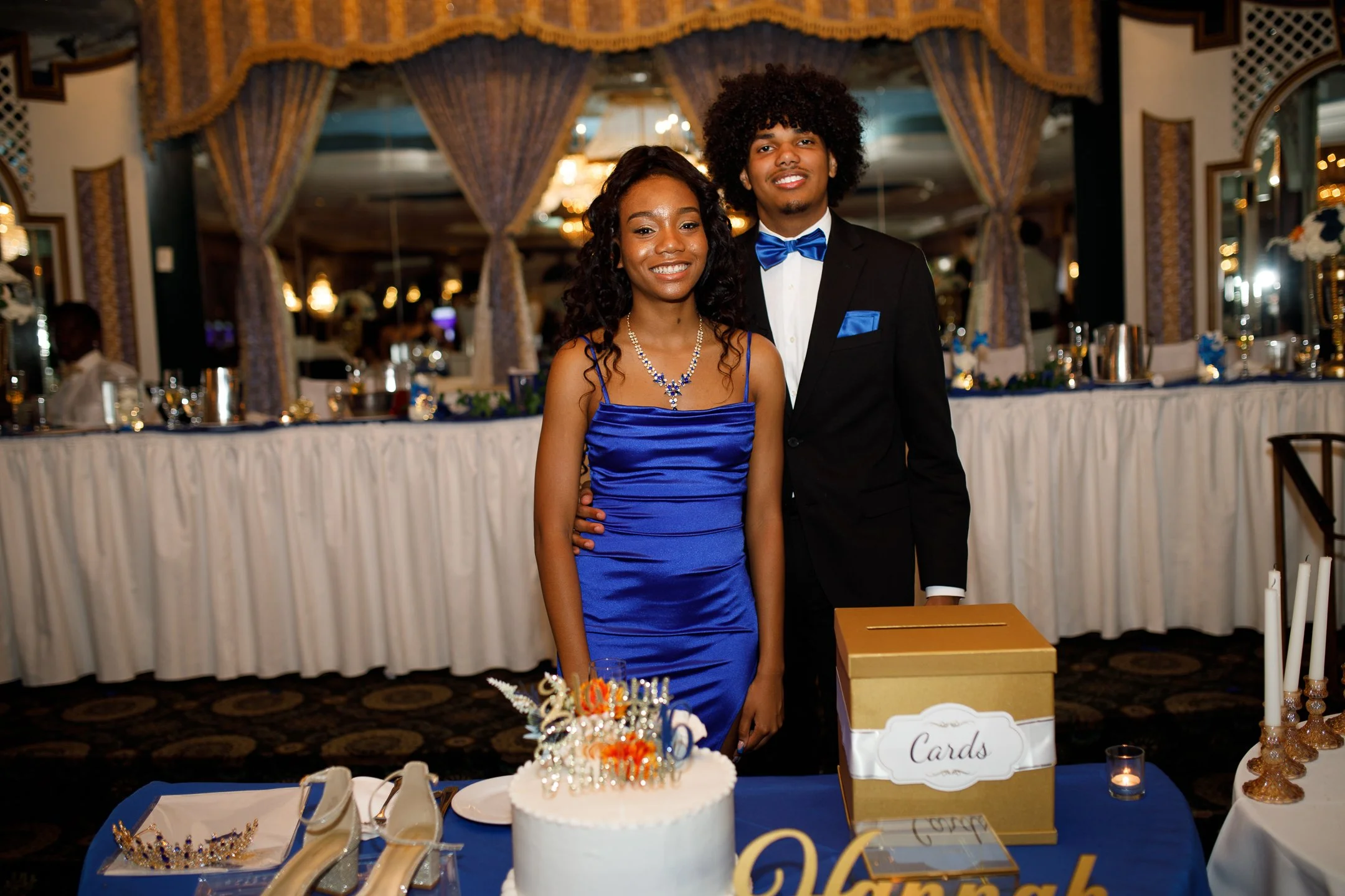 A young couple dressed in formal attire standing together at a celebratory event, with a decorated cake and a box for cards in front of them.