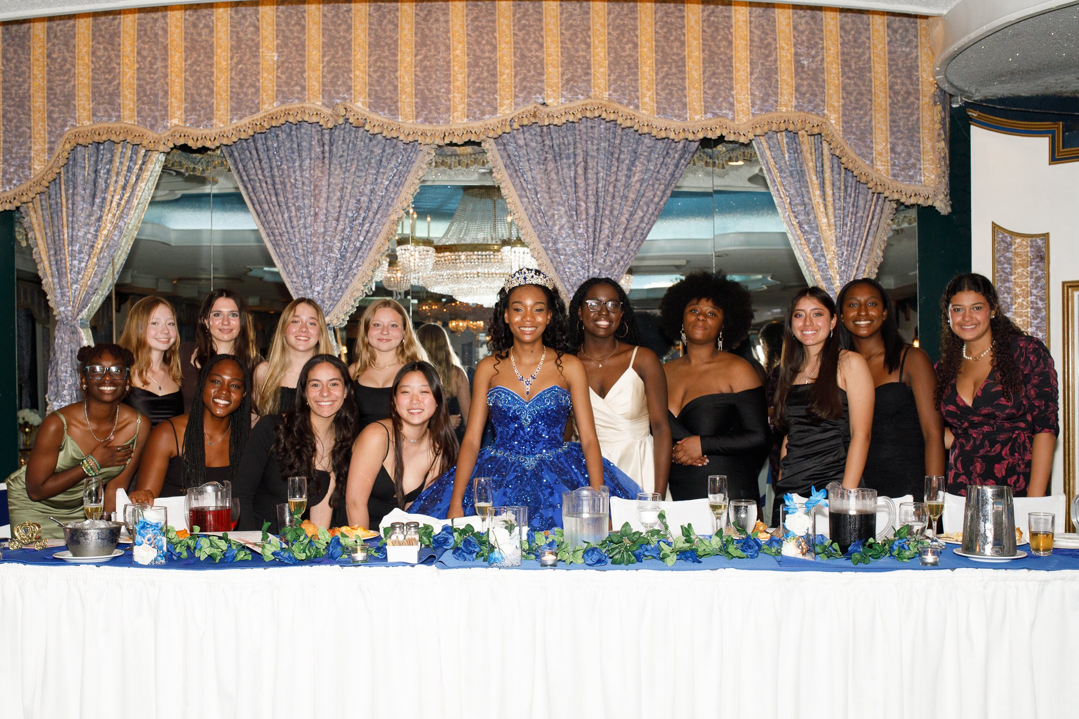 Group of young women at a formal event, some wearing dresses, standing around a decorated table in a banquet hall.